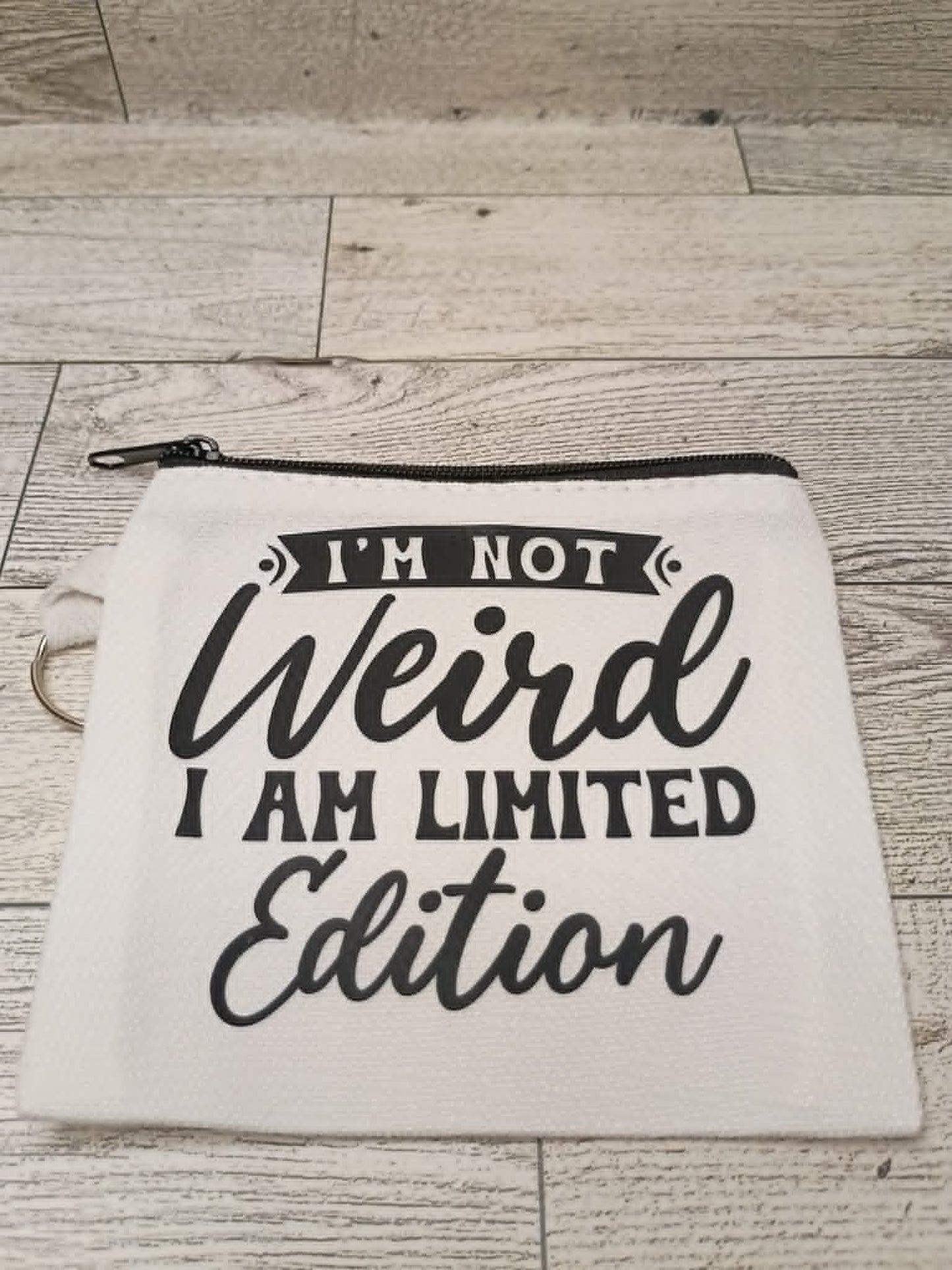 A white canvas zippered pouch with black text reading 'I'M NOT Weird I AM LIMITED Edition' laid flat on a light wood plank surface.