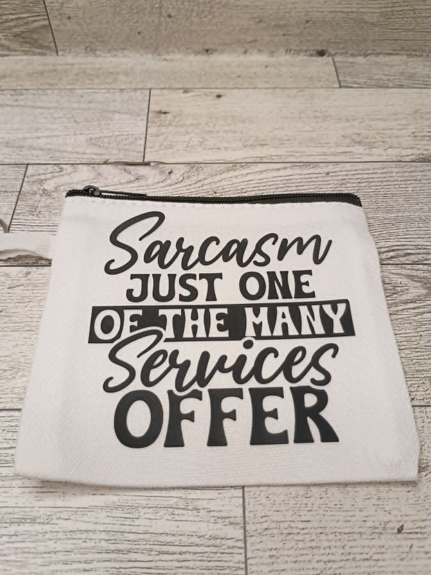 A white canvas zipper pouch with black text reading 'Sarcasm JUST ONE OF THE MANY Services OFFER' placed on a light wood plank surface.
