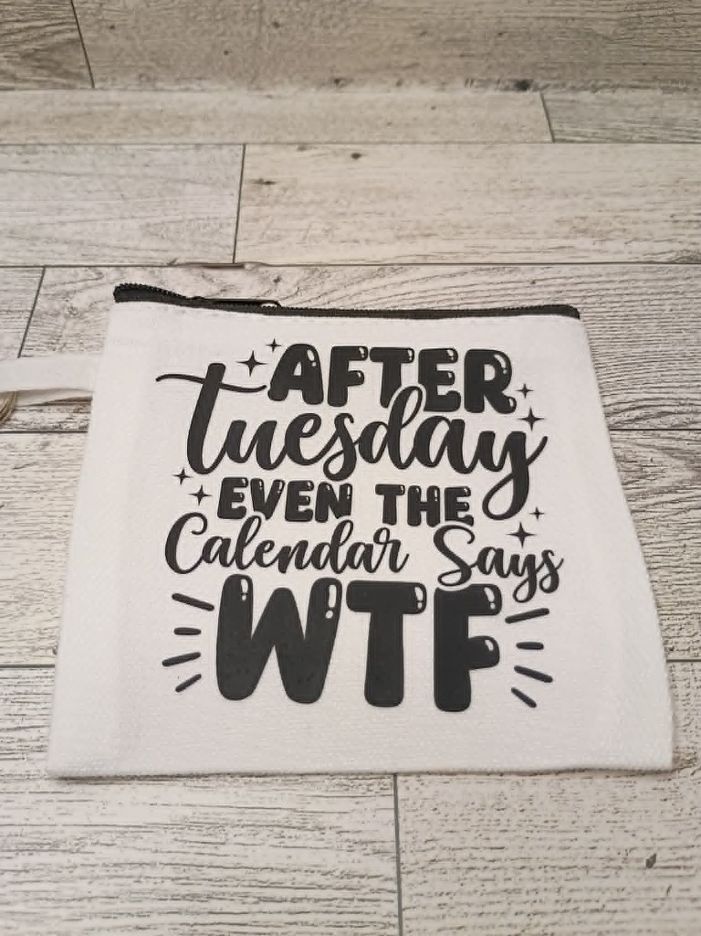 A white fabric zippered pouch with black printed text reading 'AFTER Tuesday EVEN THE Calendar Says WTF' laid flat on a light wood plank surface.