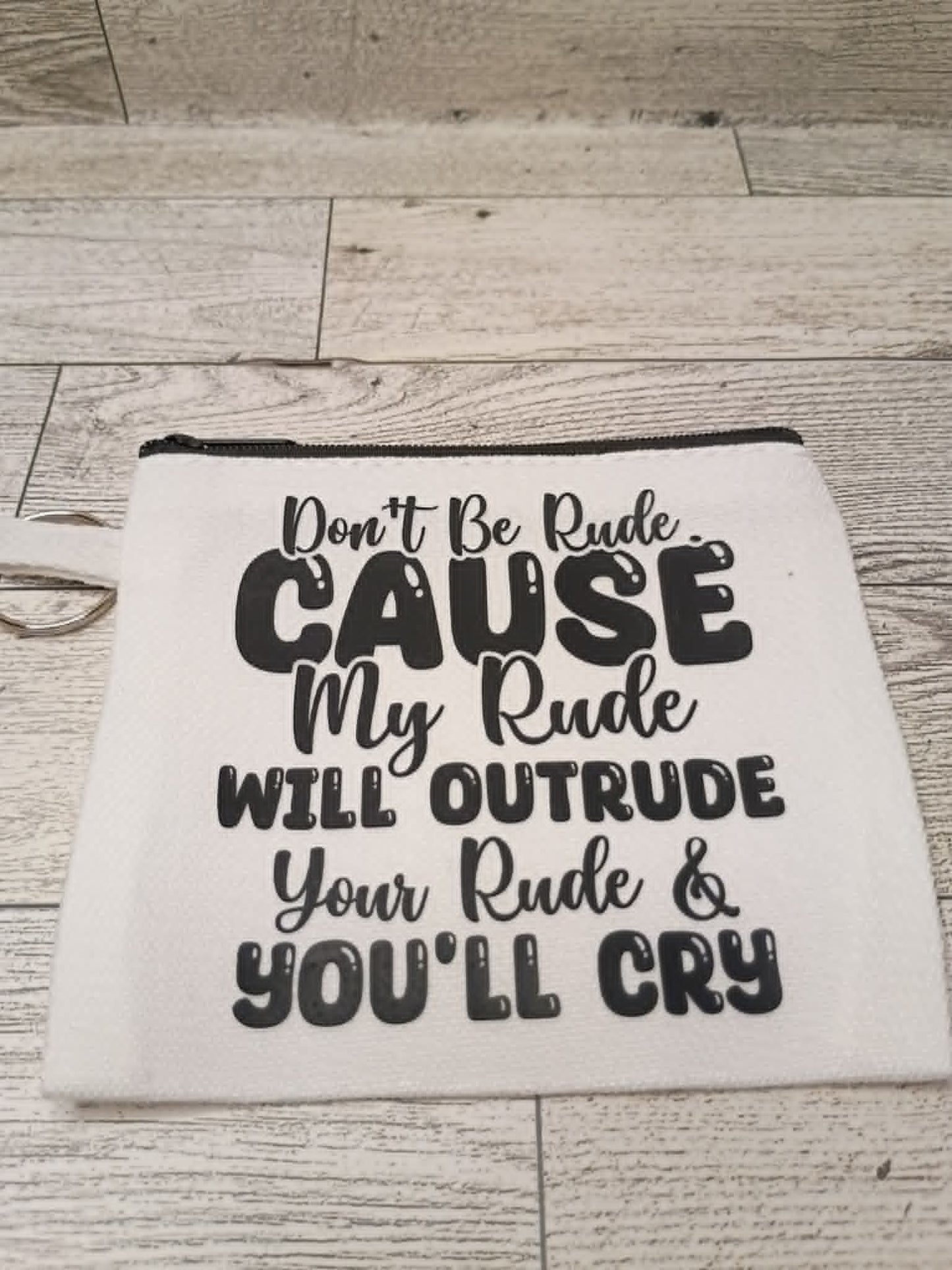 An off-white fabric pouch with a black zipper and black printed text that reads, 'Don't Be Rude. CAUSE My Rude WILL OUTRUDE Your Rude & YOU'LL CRY,' displayed on a light wood plank background.
