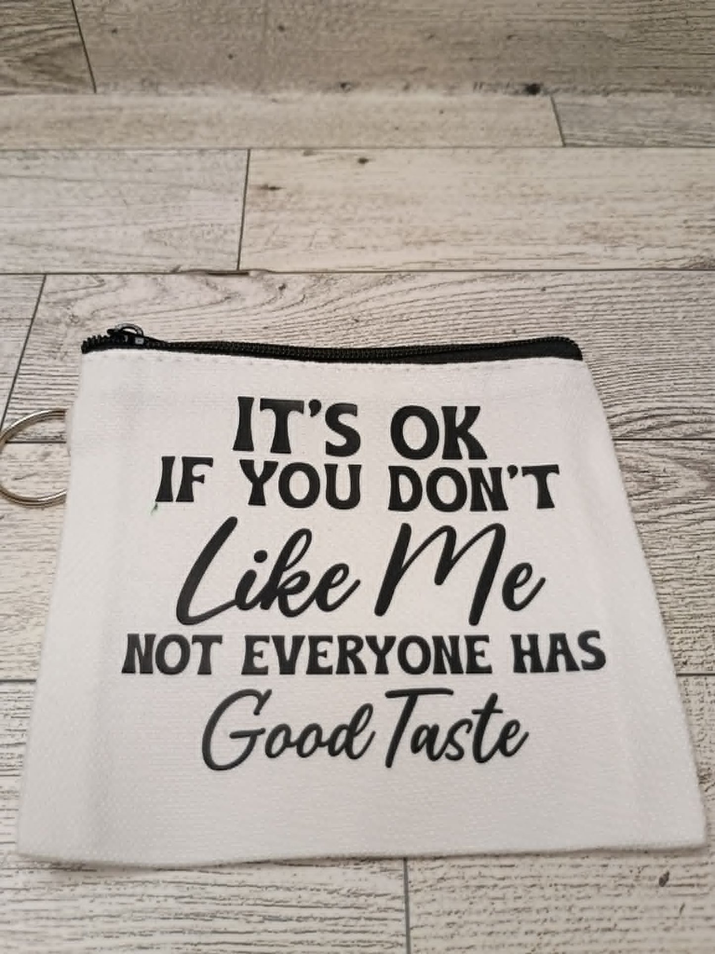 A white fabric zipper pouch with black printed text that reads, 'IT'S OK IF YOU DON'T Like Me NOT EVERYONE HAS Good Taste,' shown on a light wood plank surface.