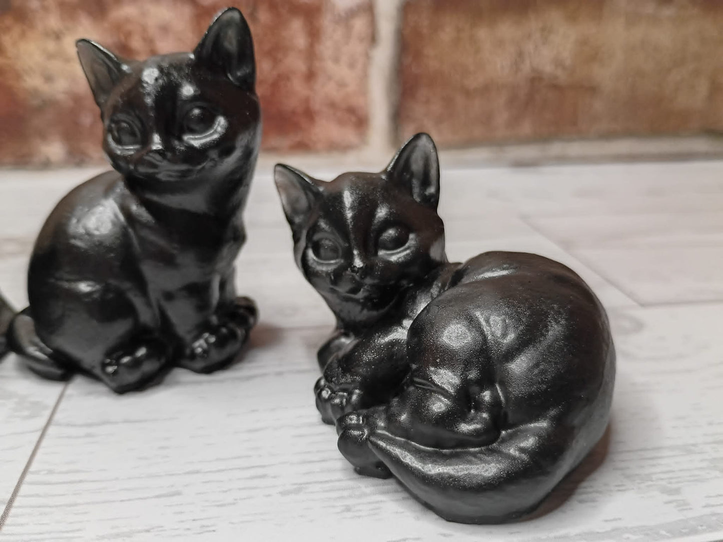 Two glossy black cat figurines, one sitting and one curled up sleeping, displayed on a light gray surface with a blurred brick wall background.