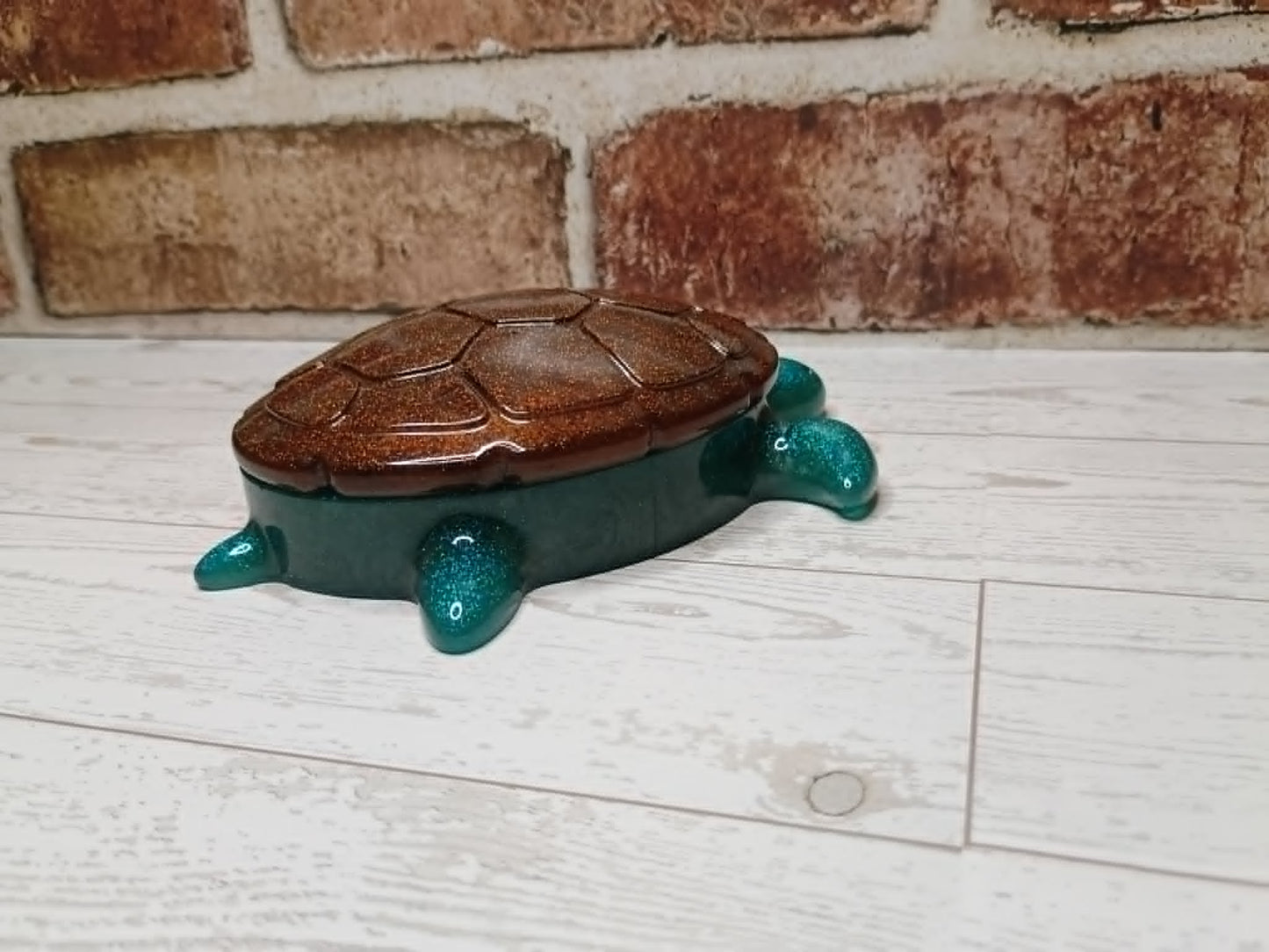 A small decorative turtle-shaped box with a brown shell and glittery teal legs and head, placed on a white wood surface in front of a brick wall.