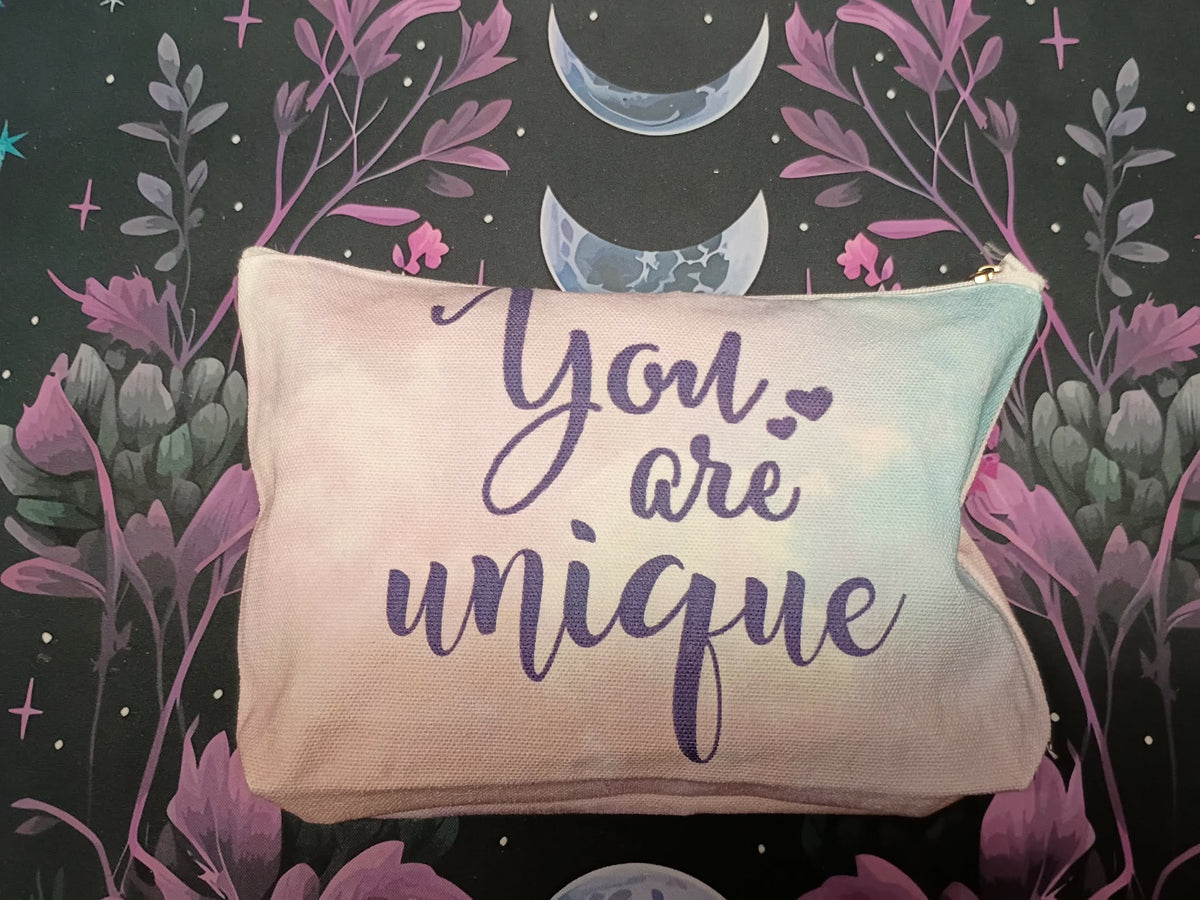 A rectangular canvas pouch with a zipper closure featuring the phrase 'You are unique' in purple script, set against a dark background with purple botanical illustrations and crescent moons.