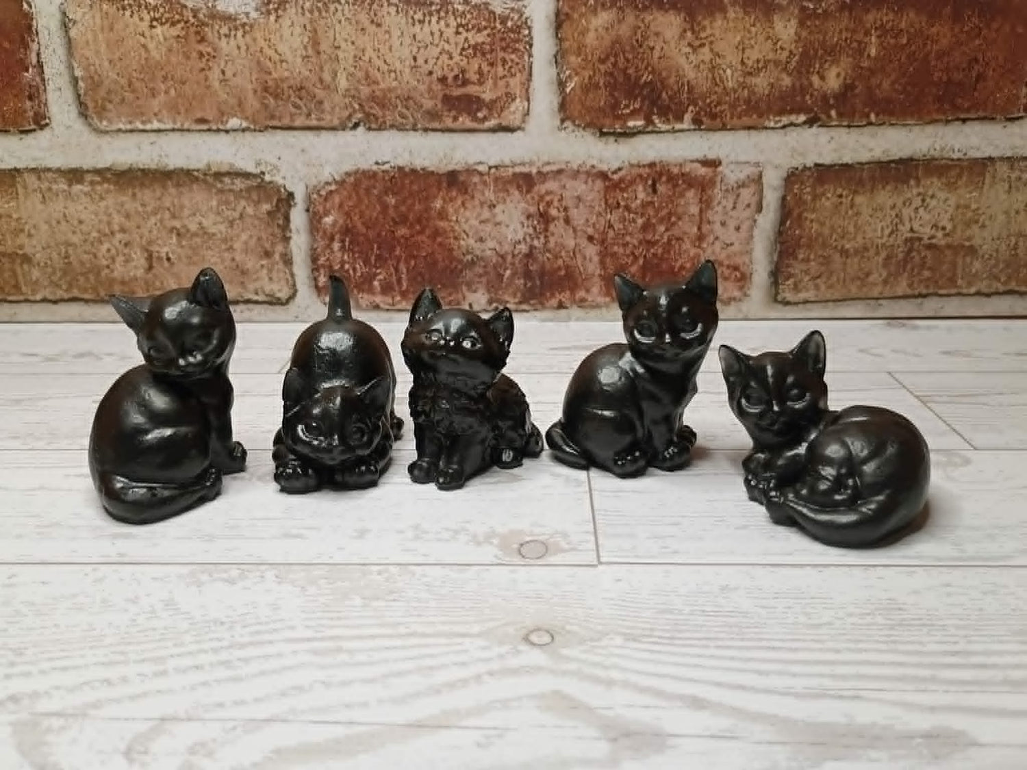 Five small black cat figurines in different poses lined up on a light wood surface in front of a brick wall.