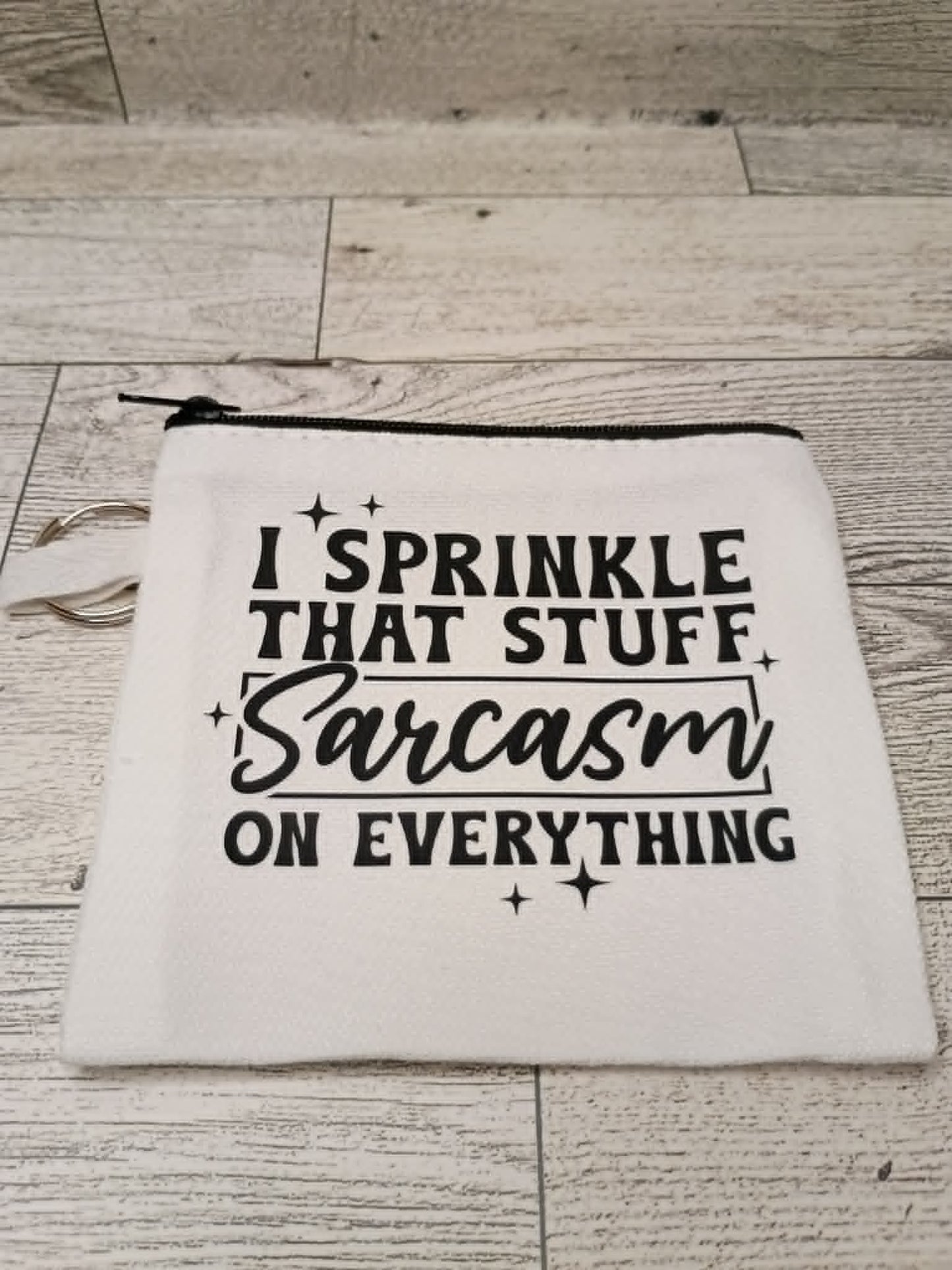A white fabric zippered pouch with black printed text reading 'I SPRINKLE THAT STUFF Sarcasm ON EVERYTHING' and small star accents, placed on a light wood floor.