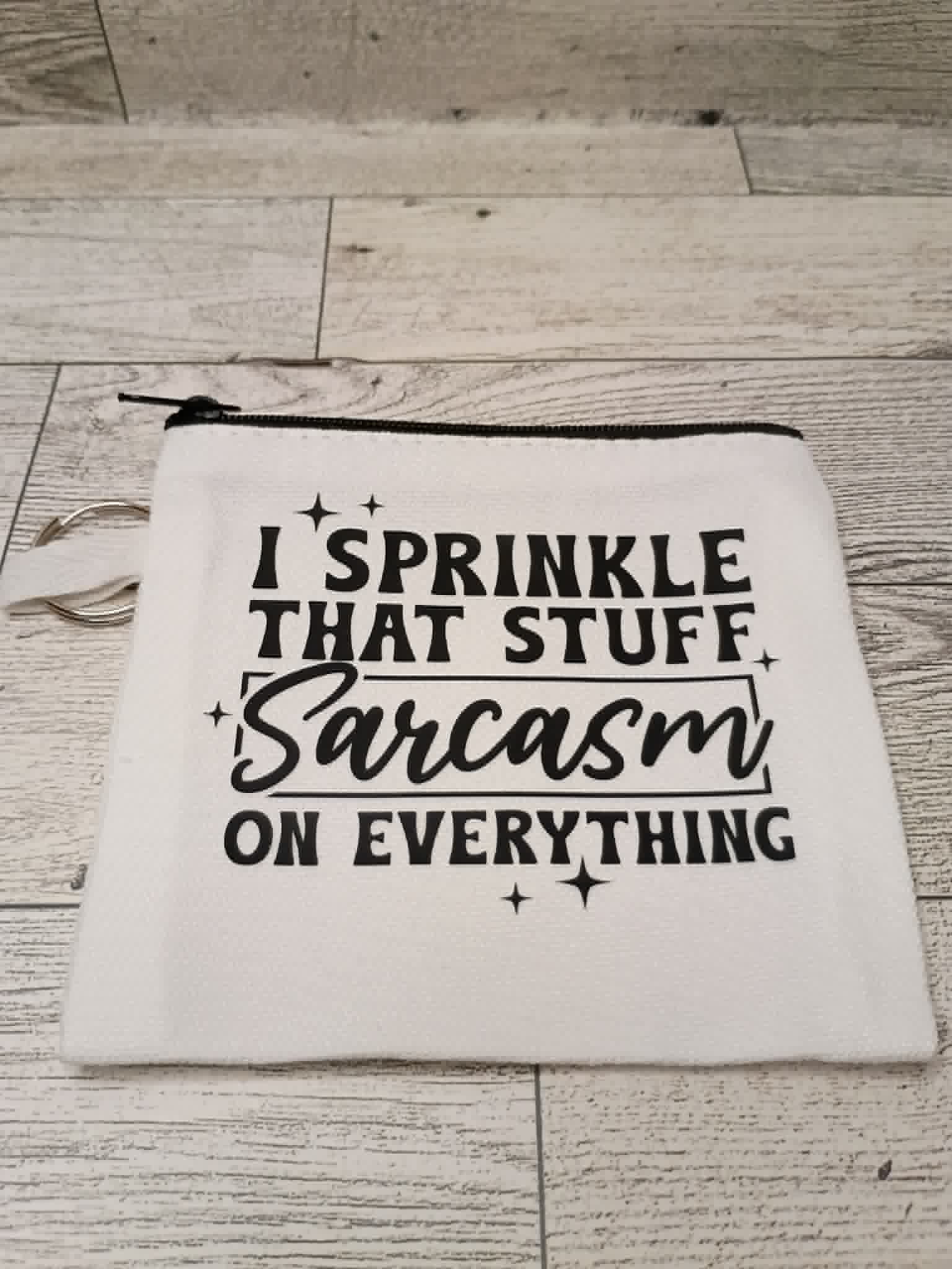A white fabric zippered pouch with black printed text reading 'I SPRINKLE THAT STUFF Sarcasm ON EVERYTHING' and small star accents, placed on a light wood floor.