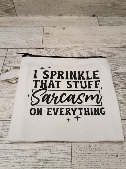 A white fabric zippered pouch with black printed text reading 'I SPRINKLE THAT STUFF Sarcasm ON EVERYTHING' and small star accents, placed on a light wood floor.
