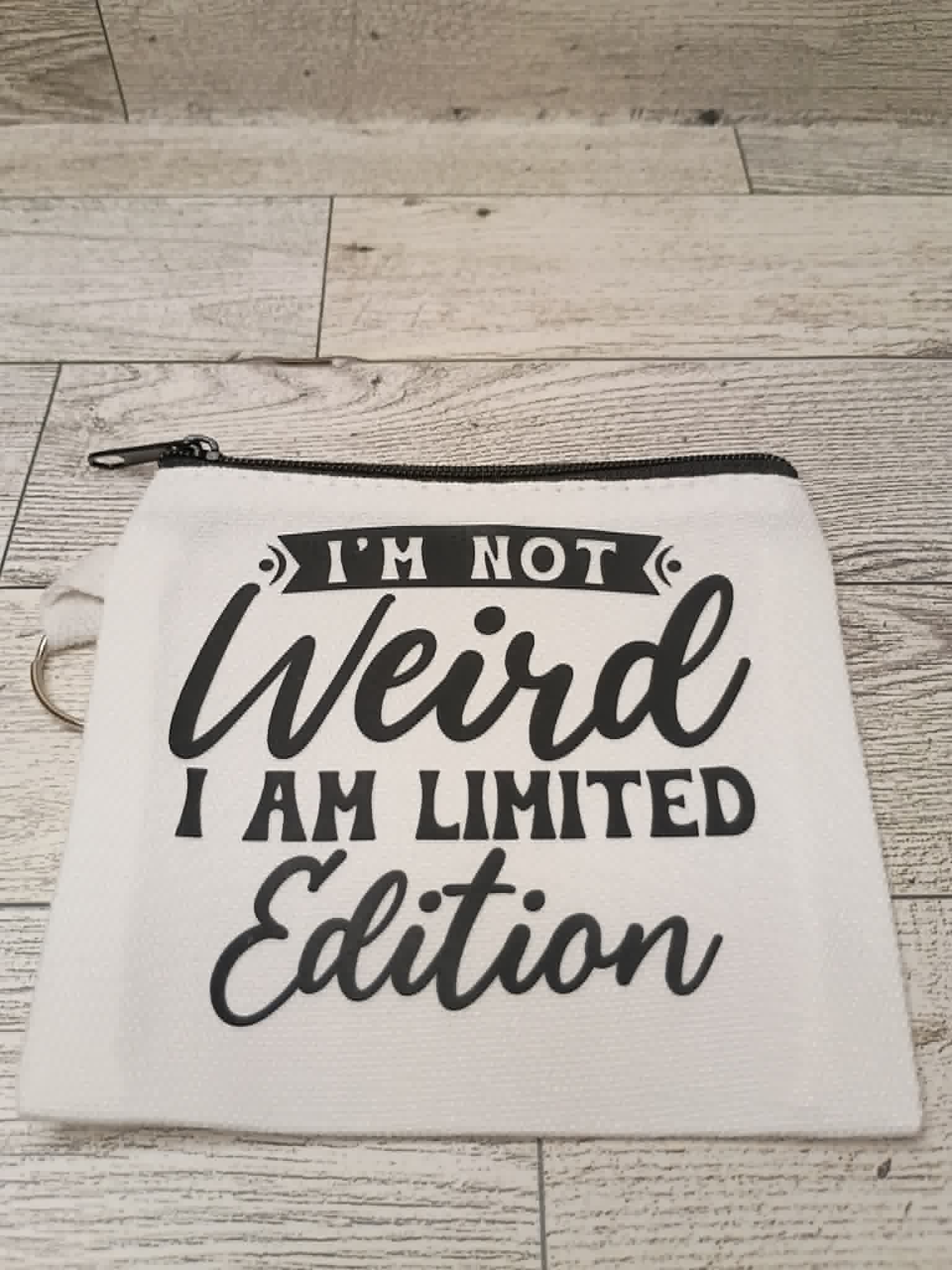 A white canvas zippered pouch with black text reading 'I'M NOT Weird I AM LIMITED Edition' laid flat on a light wood plank surface.