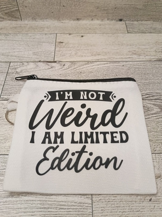 A white canvas zippered pouch with black text reading 'I'M NOT Weird I AM LIMITED Edition' laid flat on a light wood plank surface.