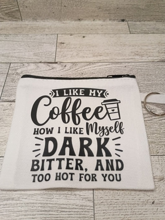 A white fabric zipper pouch with black printed text that reads 'I LIKE MY Coffee HOW I LIKE Myself DARK BITTER, AND TOO HOT FOR YOU,' shown on a light wood floor with a metal keyring nearby.