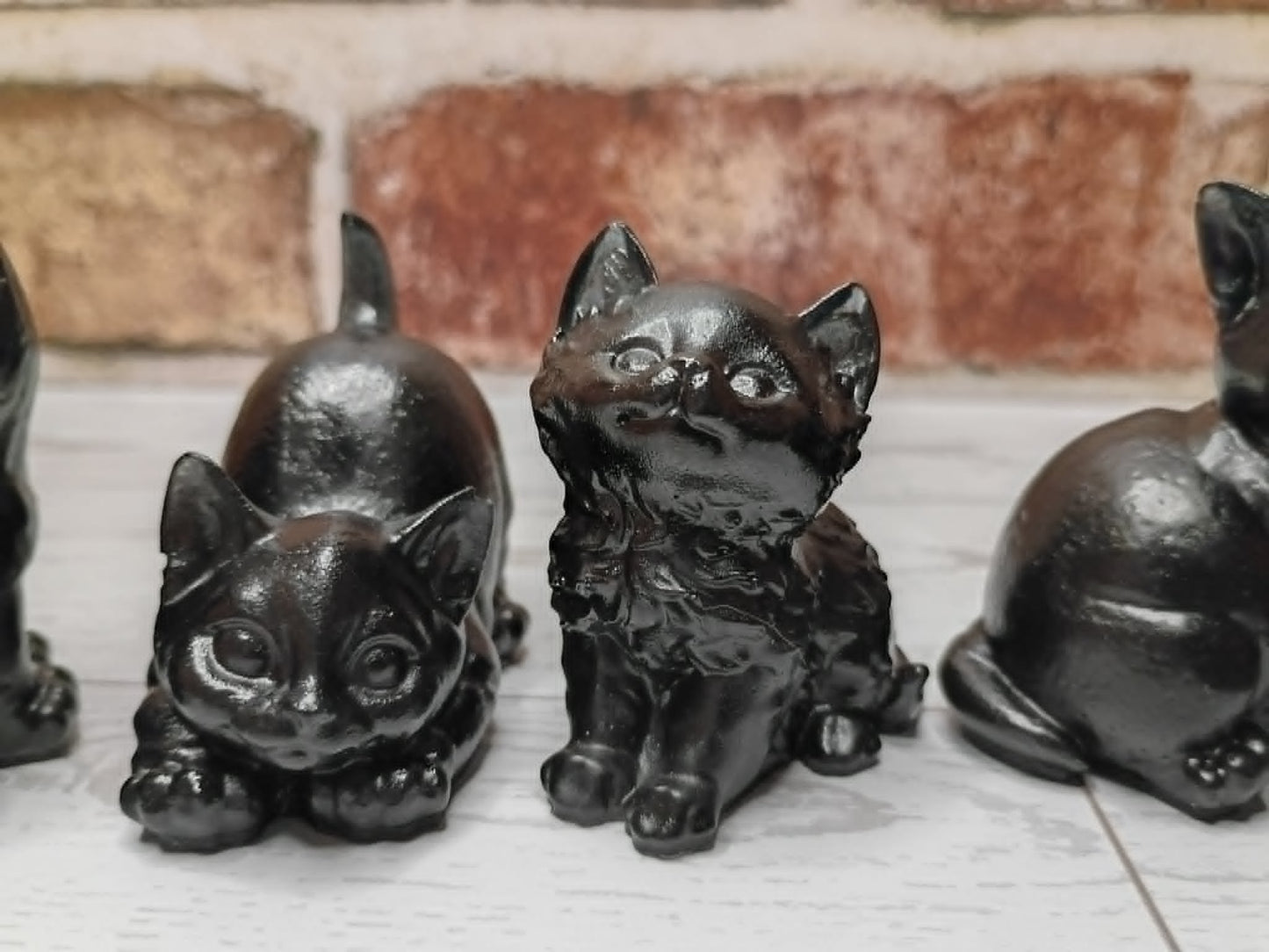 A set of glossy black cast-iron cat figurines in various poses, displayed on a white wooden surface with a rustic brick wall background.