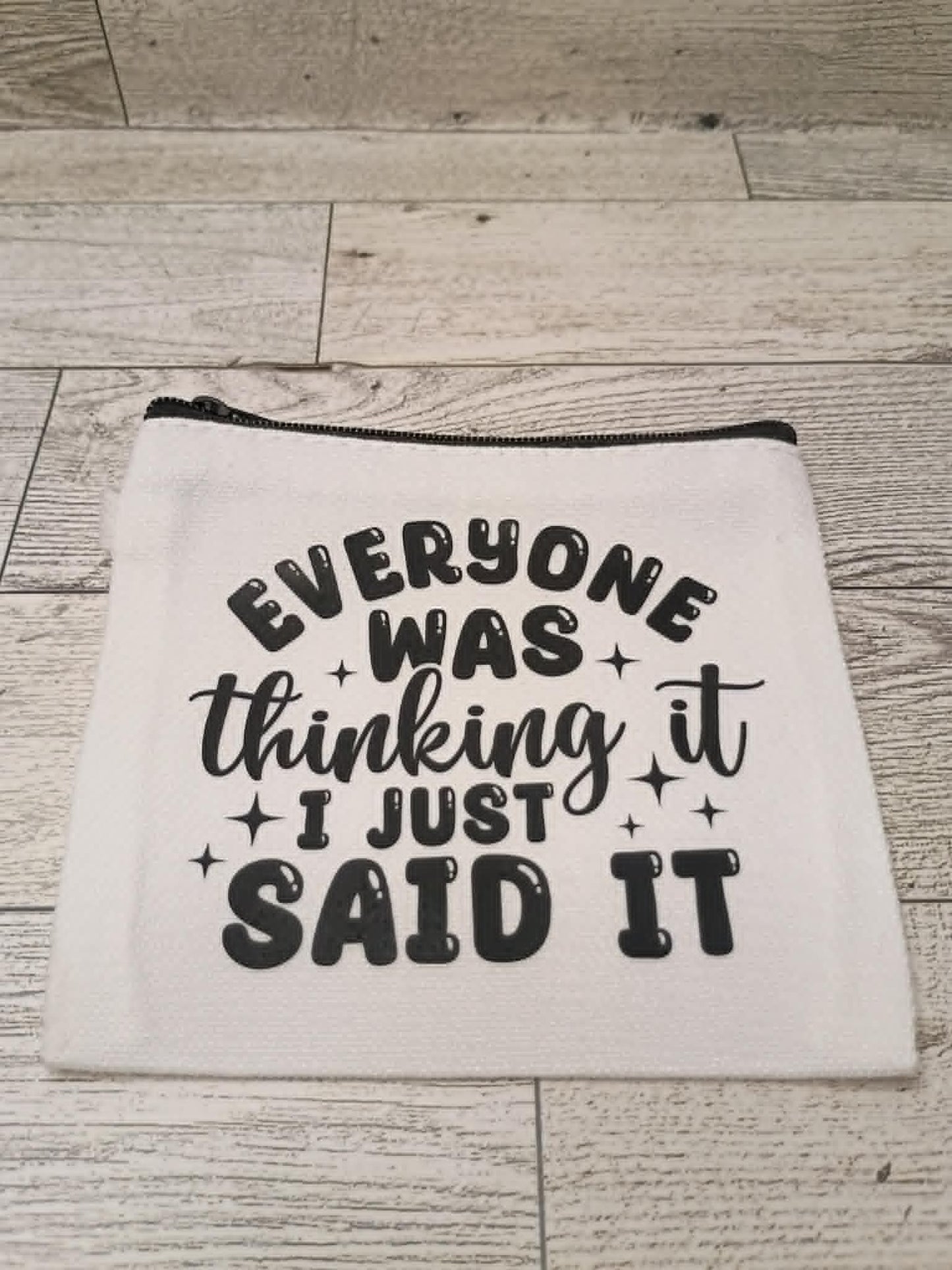 A white fabric zippered pouch with black text reading 'EVERYONE WAS thinking it I JUST SAID IT' and small star accents, placed on a light wood plank surface.