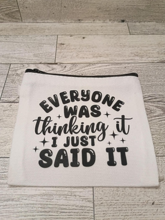 A white fabric zippered pouch with black text reading 'EVERYONE WAS thinking it I JUST SAID IT' and small star accents, placed on a light wood plank surface.