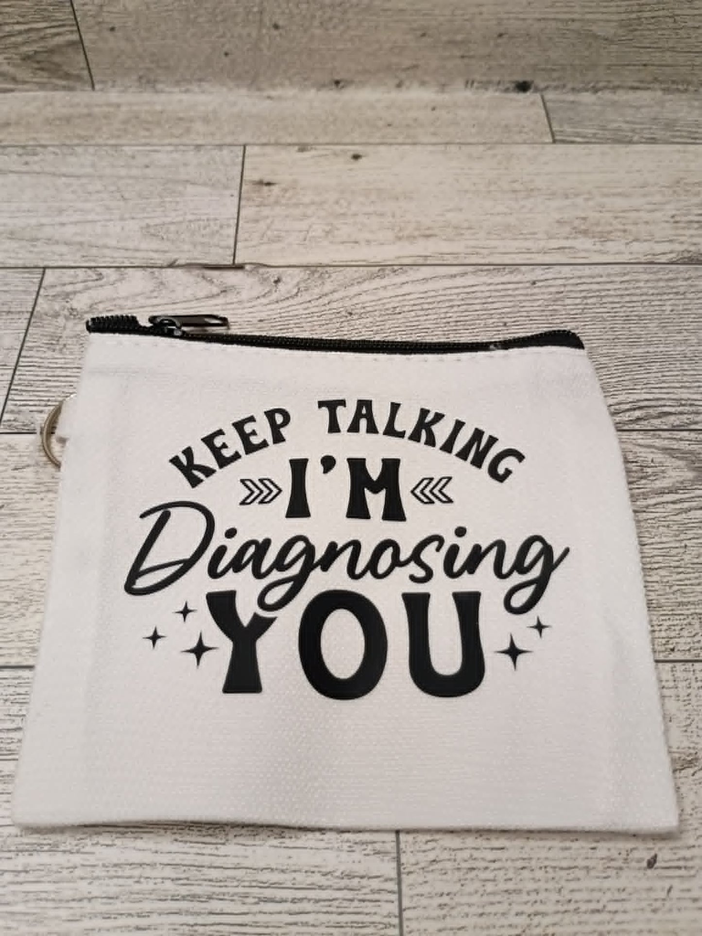 A white fabric zippered pouch with black text reading 'KEEP TALKING I'M Diagnosing YOU' and small star accents, placed on a light wood floor.