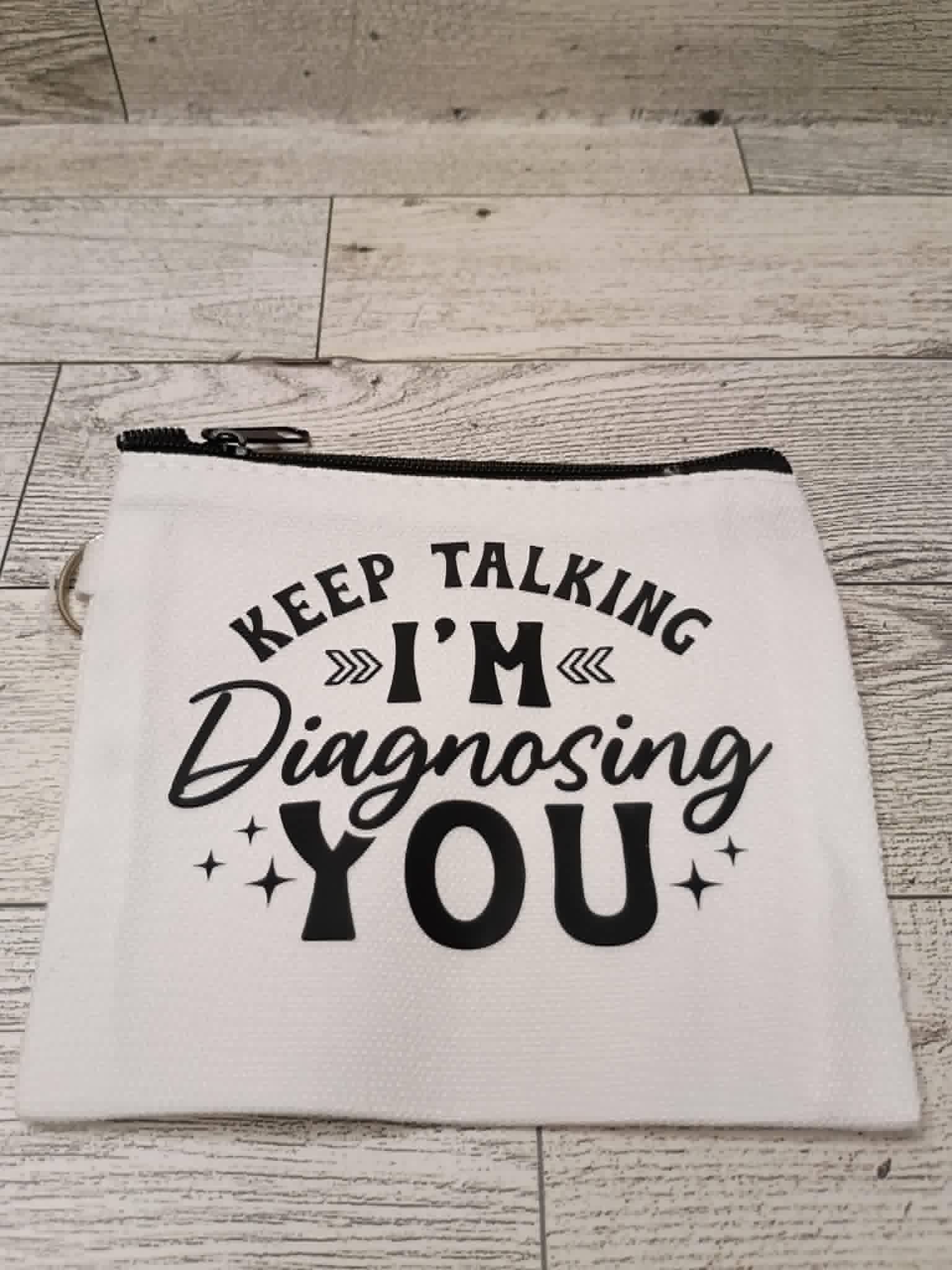 A white fabric zippered pouch with black text reading 'KEEP TALKING I'M Diagnosing YOU' and small star accents, placed on a light wood floor.