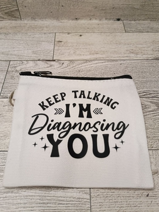 A white fabric zippered pouch with black text reading 'KEEP TALKING I'M Diagnosing YOU' and small star accents, placed on a light wood floor.