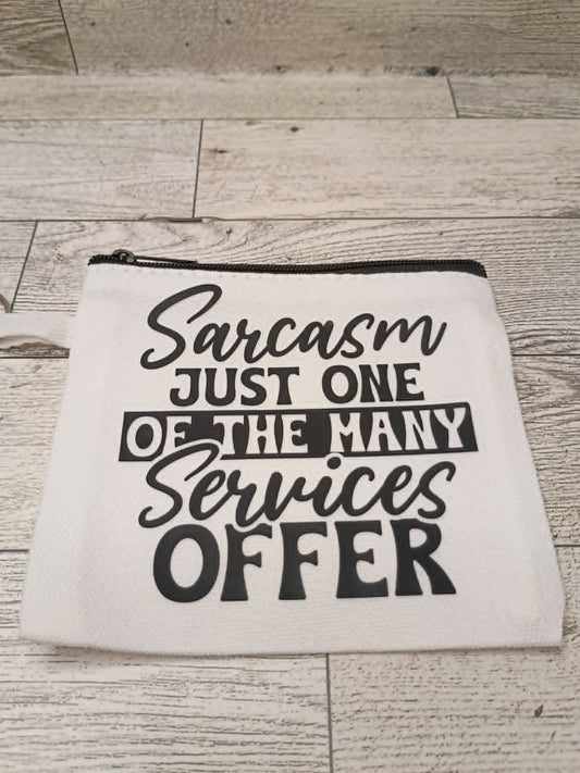 A white canvas zipper pouch with black text reading 'Sarcasm JUST ONE OF THE MANY Services OFFER' placed on a light wood plank surface.
