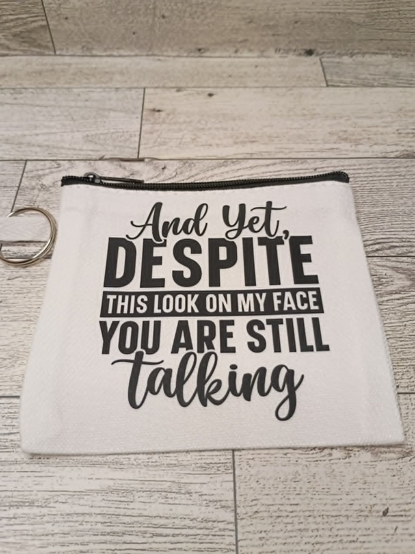 A white fabric cosmetic pouch with a black zipper and a metal key ring, printed with the humorous phrase 'And Yet, DESPITE THIS LOOK ON MY FACE YOU ARE STILL talking' in black lettering, displayed on a light gray wood plank background.