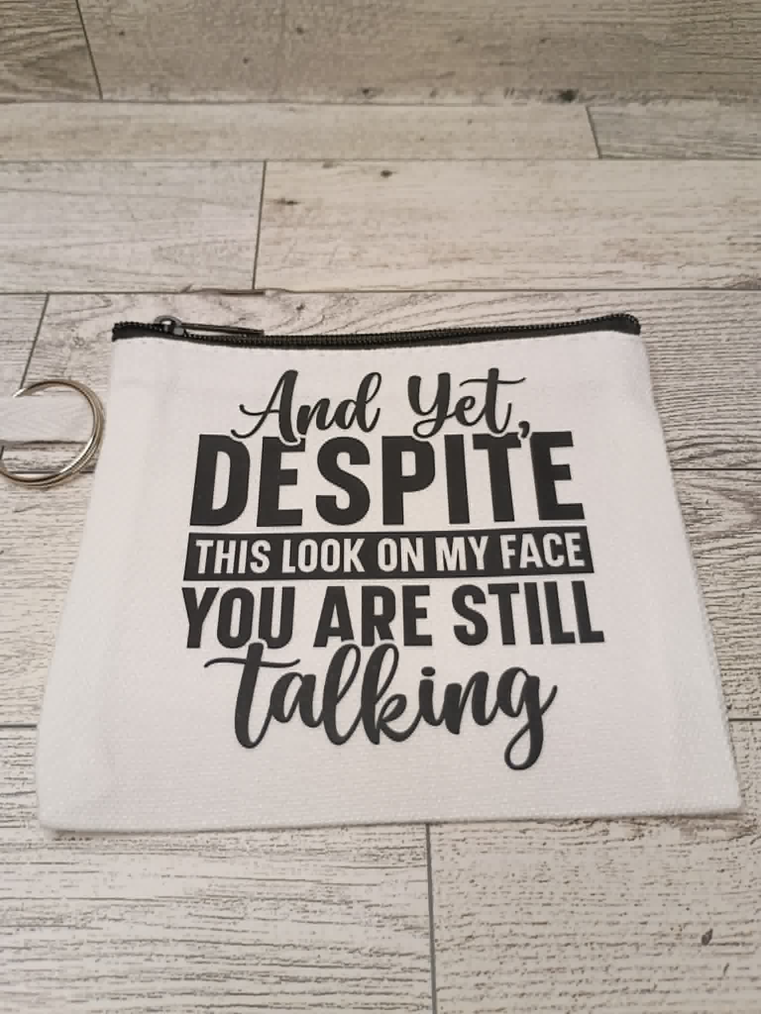 A white fabric cosmetic pouch with a black zipper and a metal key ring, printed with the humorous phrase 'And Yet, DESPITE THIS LOOK ON MY FACE YOU ARE STILL talking' in black lettering, displayed on a light gray wood plank background.