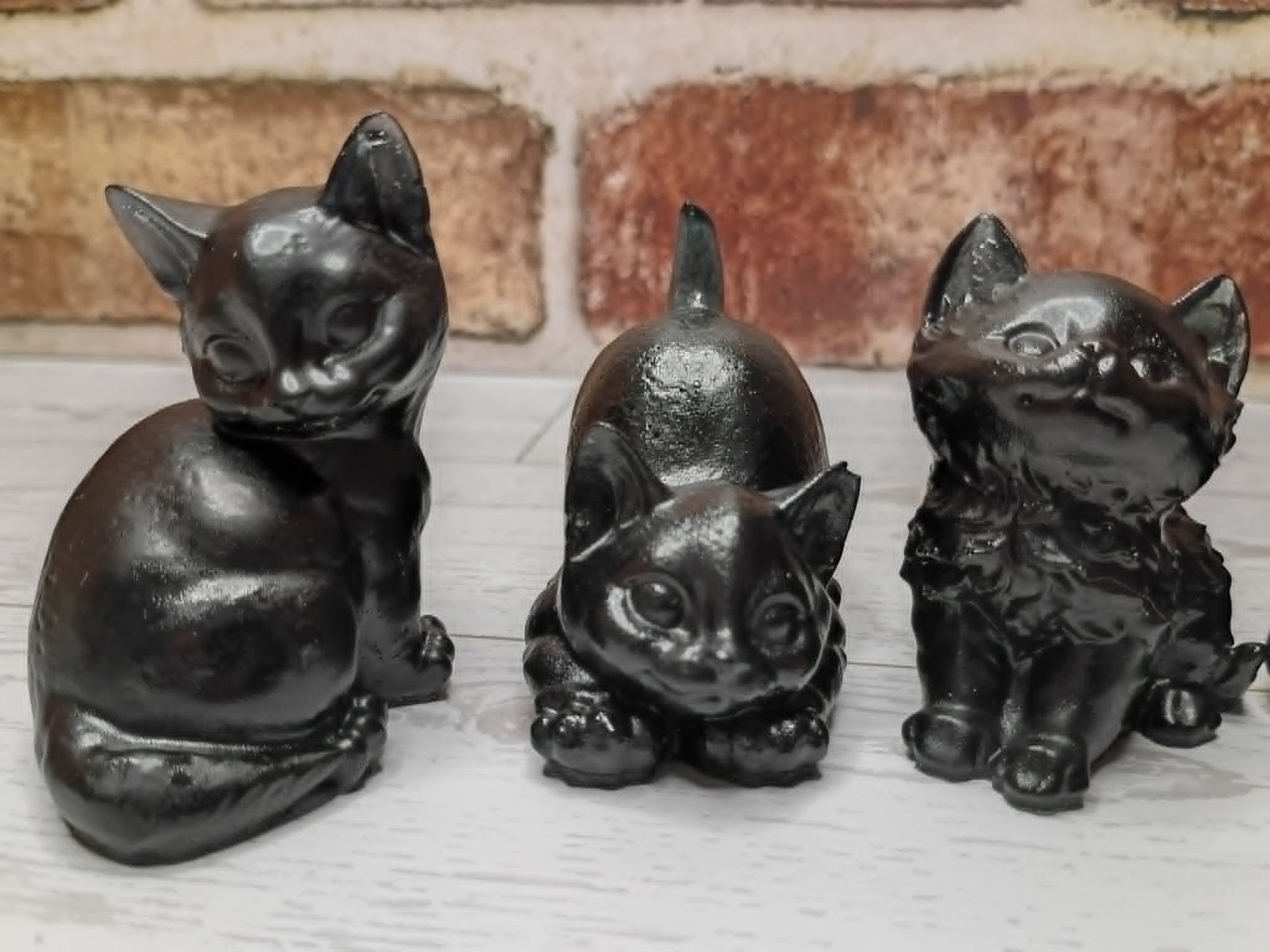 Three small black cat figurines in different poses, displayed on a light surface with a rustic brick wall background.