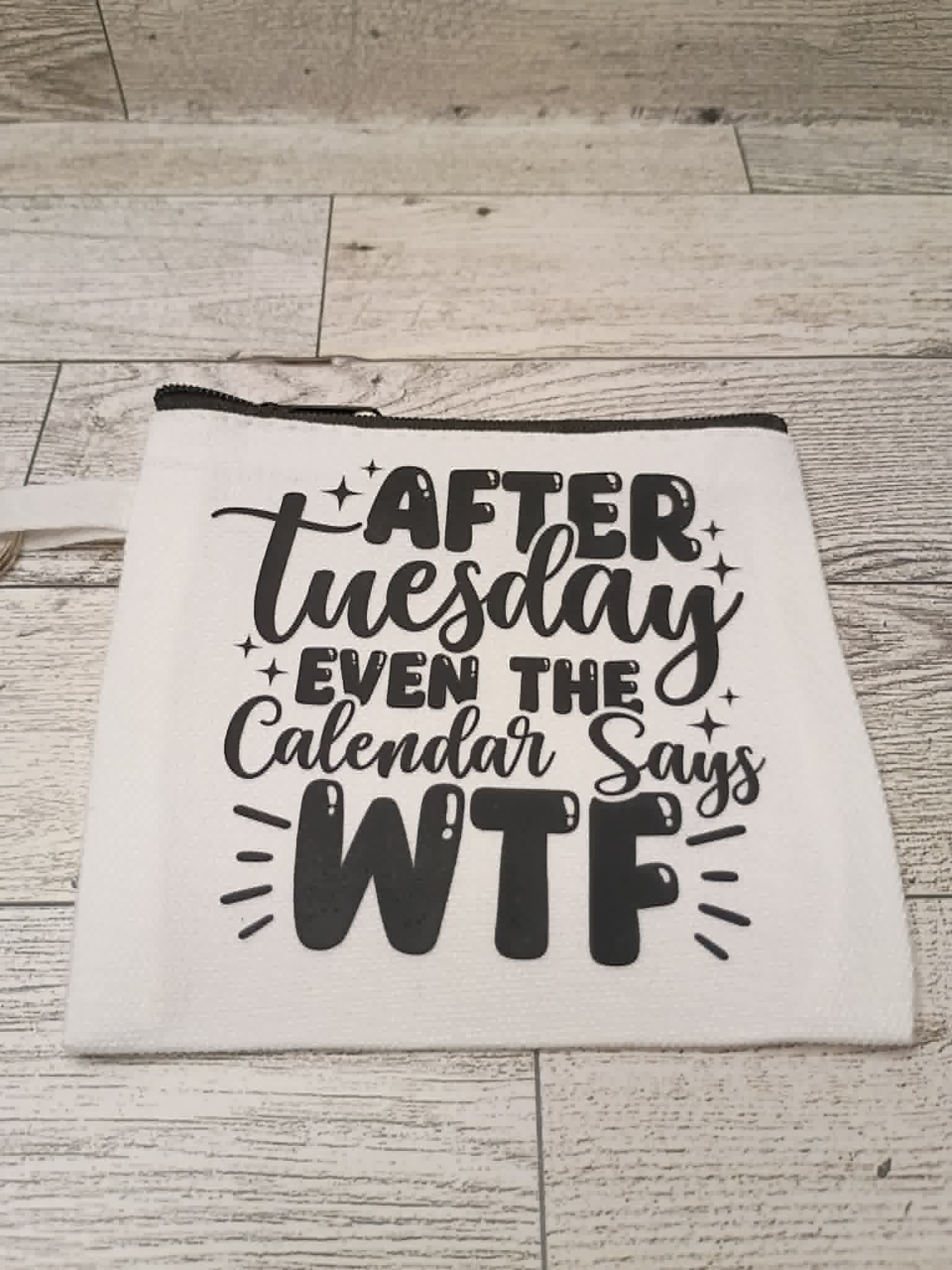 A white fabric zippered pouch with black printed text reading 'AFTER Tuesday EVEN THE Calendar Says WTF' laid flat on a light wood plank surface.