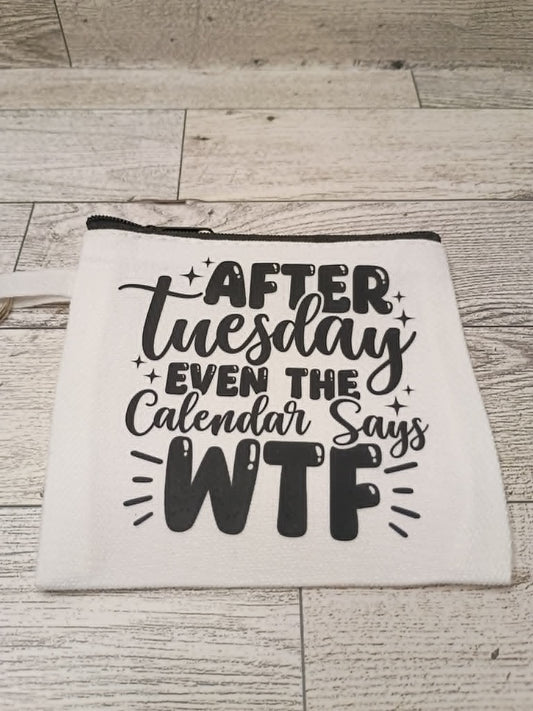 A white fabric zippered pouch with black printed text reading 'AFTER Tuesday EVEN THE Calendar Says WTF' laid flat on a light wood plank surface.