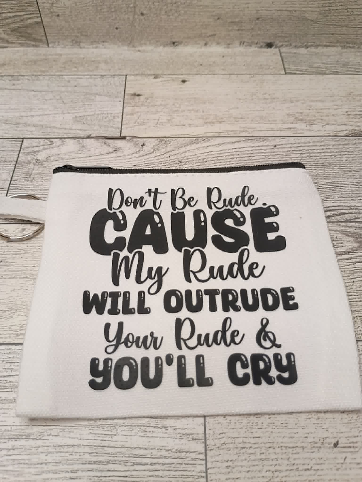 An off-white fabric pouch with a black zipper and black printed text that reads, 'Don't Be Rude. CAUSE My Rude WILL OUTRUDE Your Rude & YOU'LL CRY,' displayed on a light wood plank background.