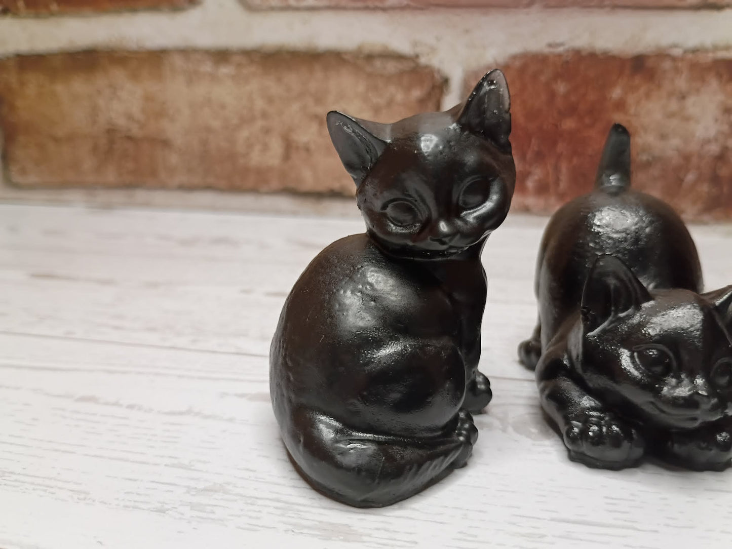 Two small black cat figurines displayed on a white wooden surface with a blurred brick wall in the background.