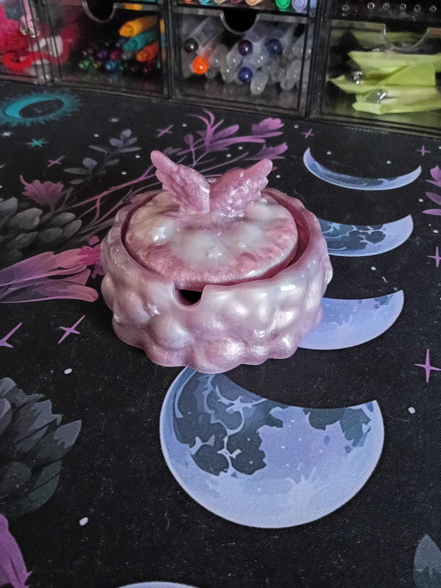 Pink ceramic teapot with butterfly wings on a dark surface with moon and star designs.