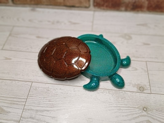 A small turtle-shaped container or dish with a glossy brown shell and a translucent teal-green body, placed on a light wood floor.