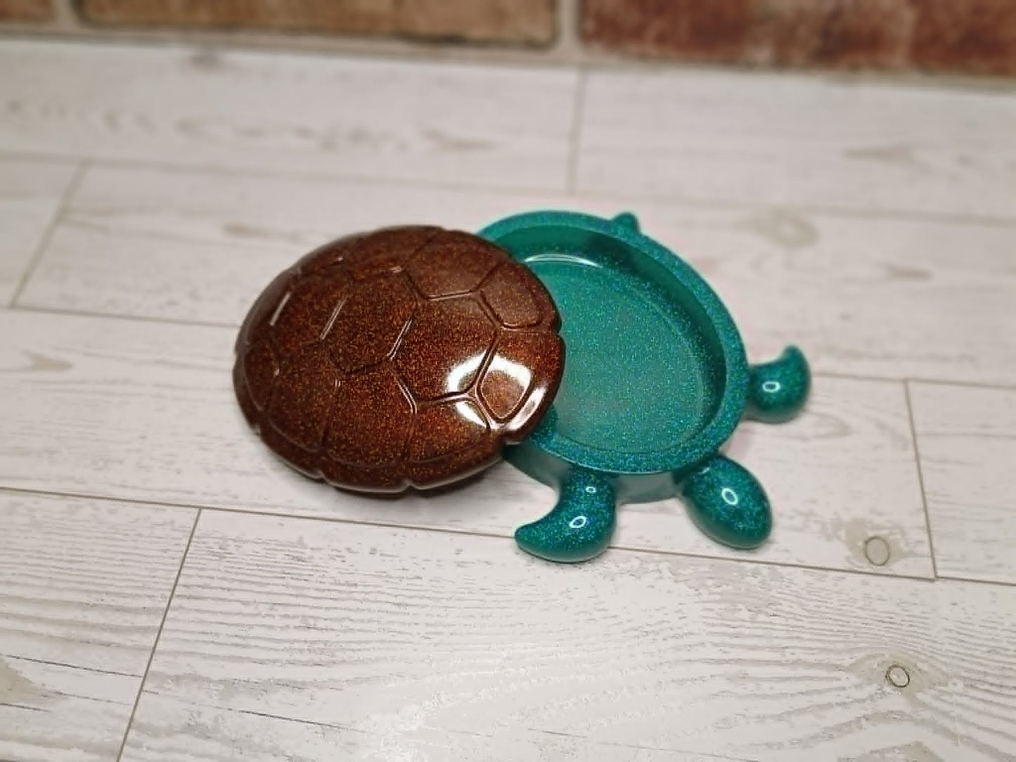 A small turtle-shaped container or dish with a glossy brown shell and a translucent teal-green body, placed on a light wood floor.