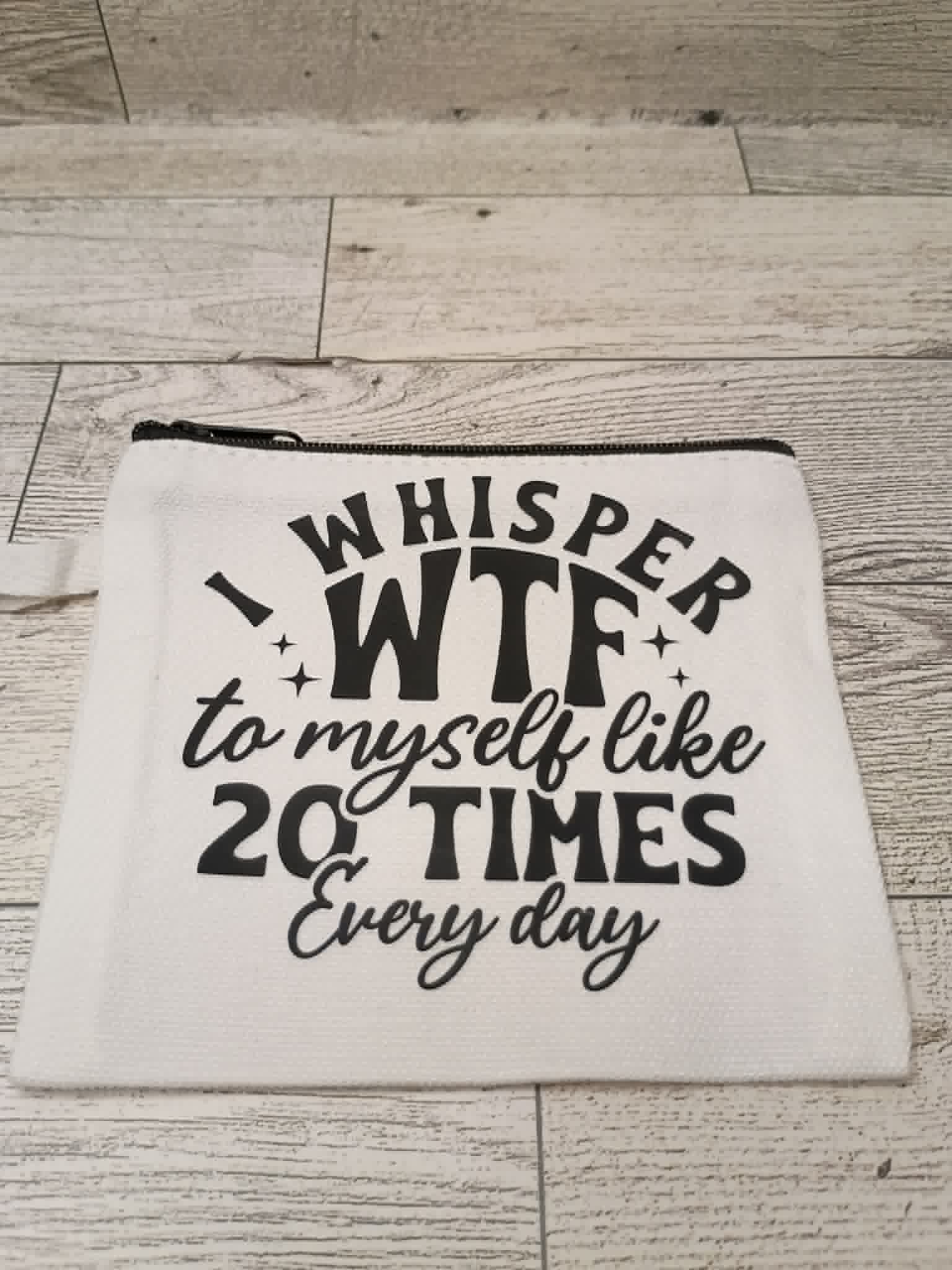 A white fabric zippered pouch with black printed text reading 'I WHISPER WTF to myself like 20 TIMES Every day', placed on a light wood plank surface.