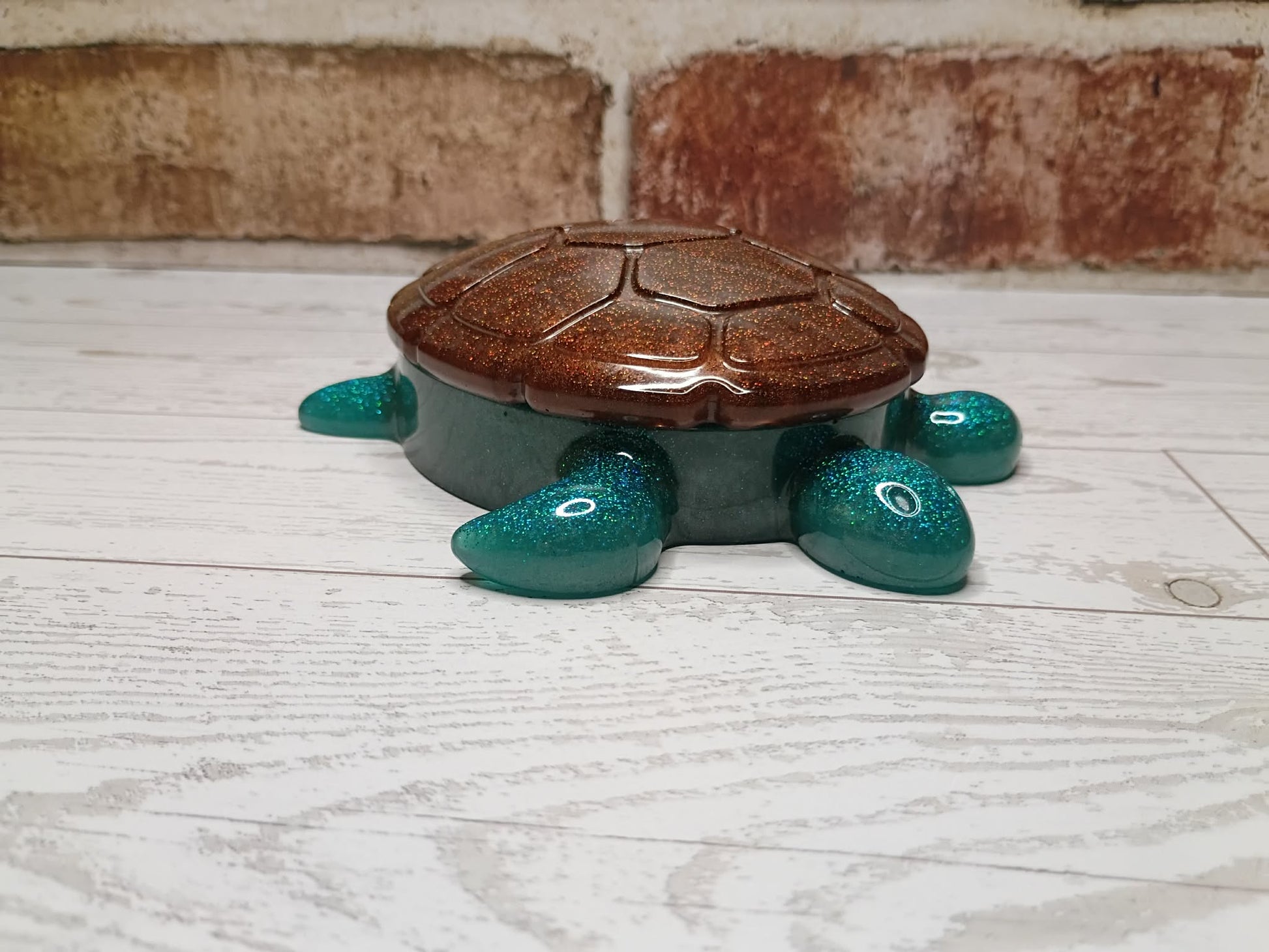 A small decorative turtle-shaped container or box with a glittery brown shell and dark teal body and legs, placed on a white wood surface with a brick wall in the background.