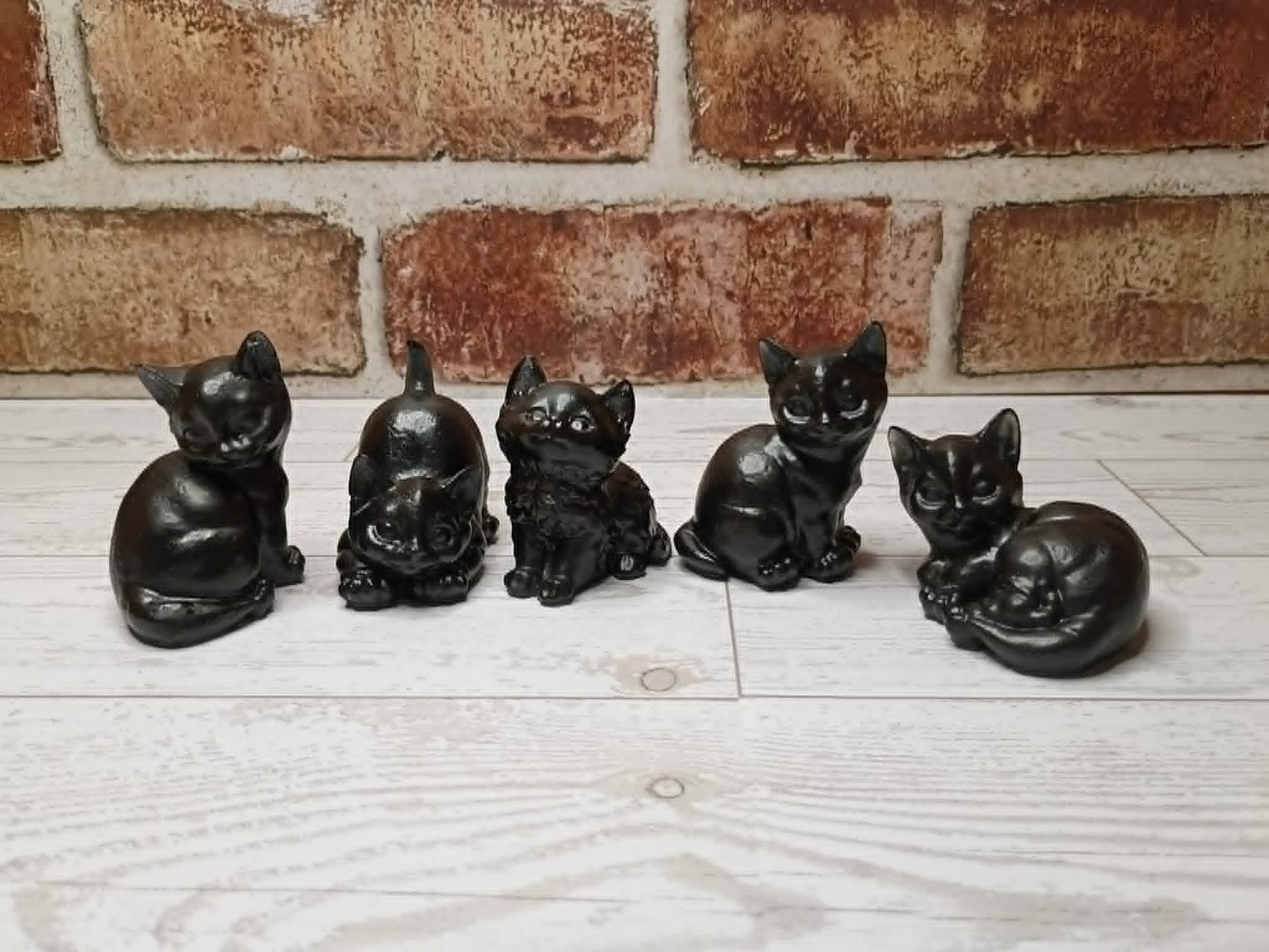 Five small black cat figurines in different poses lined up on a light wood surface in front of a brick wall.