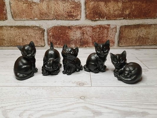 Five small black cat figurines in different poses lined up on a light wood surface in front of a brick wall.