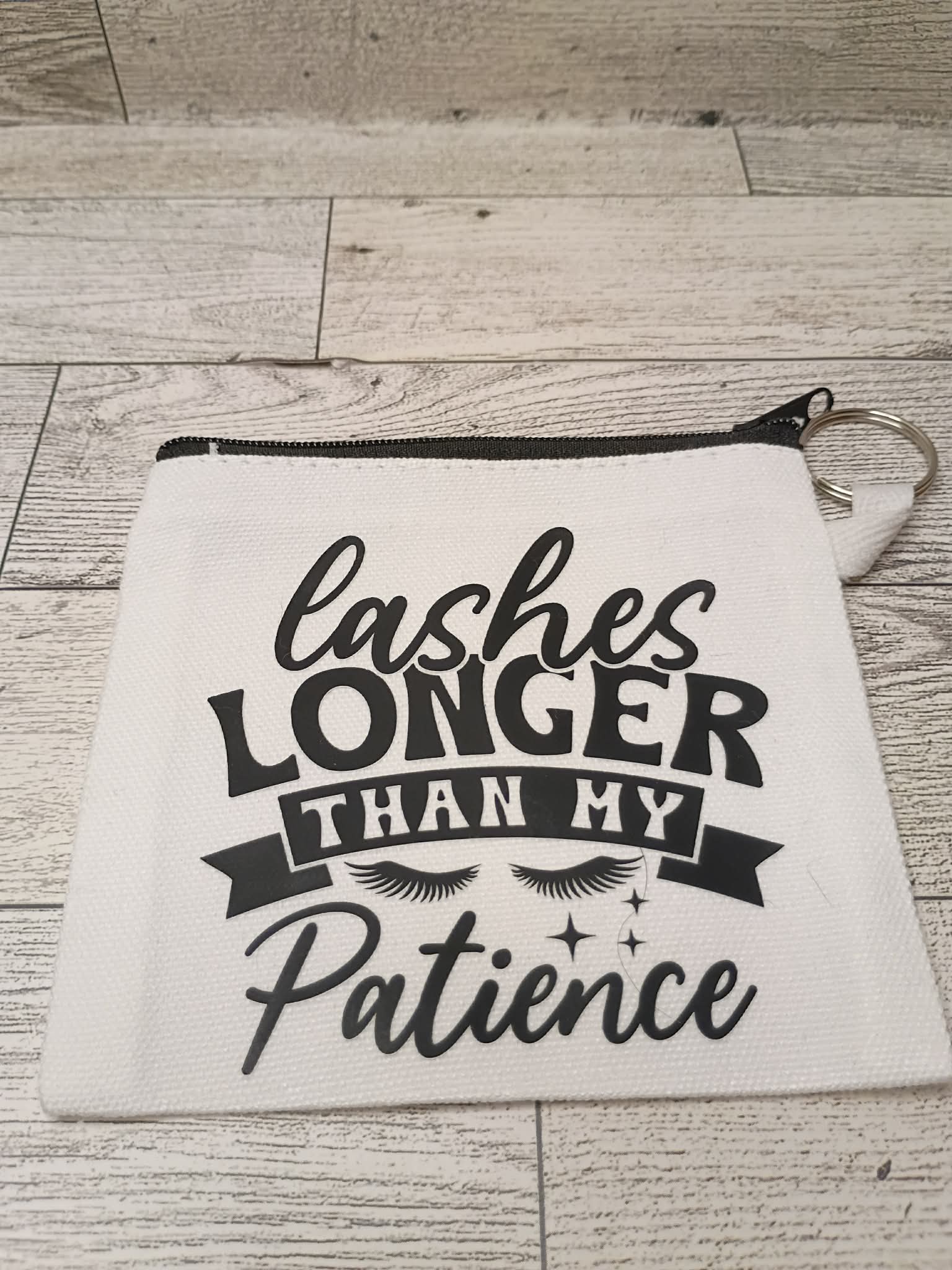 A white canvas cosmetic pouch with a black zipper and a printed black graphic that reads 'lashes LONGER THAN MY Patience' with eyelashes and stars, placed on a light wood plank surface.