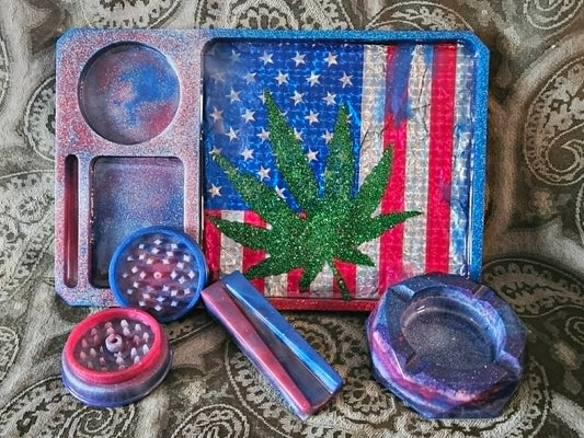 Set of cannabis accessories including a tray with a marijuana leaf design, grinder, and ashtray on a patterned surface.