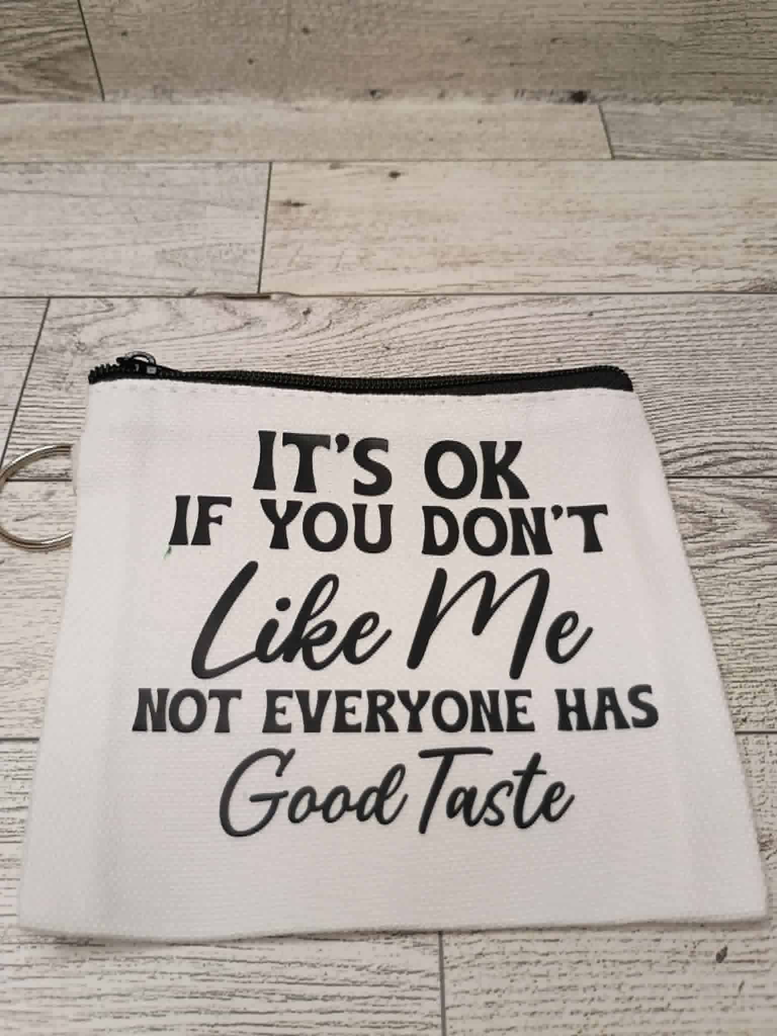 A white fabric zipper pouch with black printed text that reads, 'IT'S OK IF YOU DON'T Like Me NOT EVERYONE HAS Good Taste,' shown on a light wood plank surface.