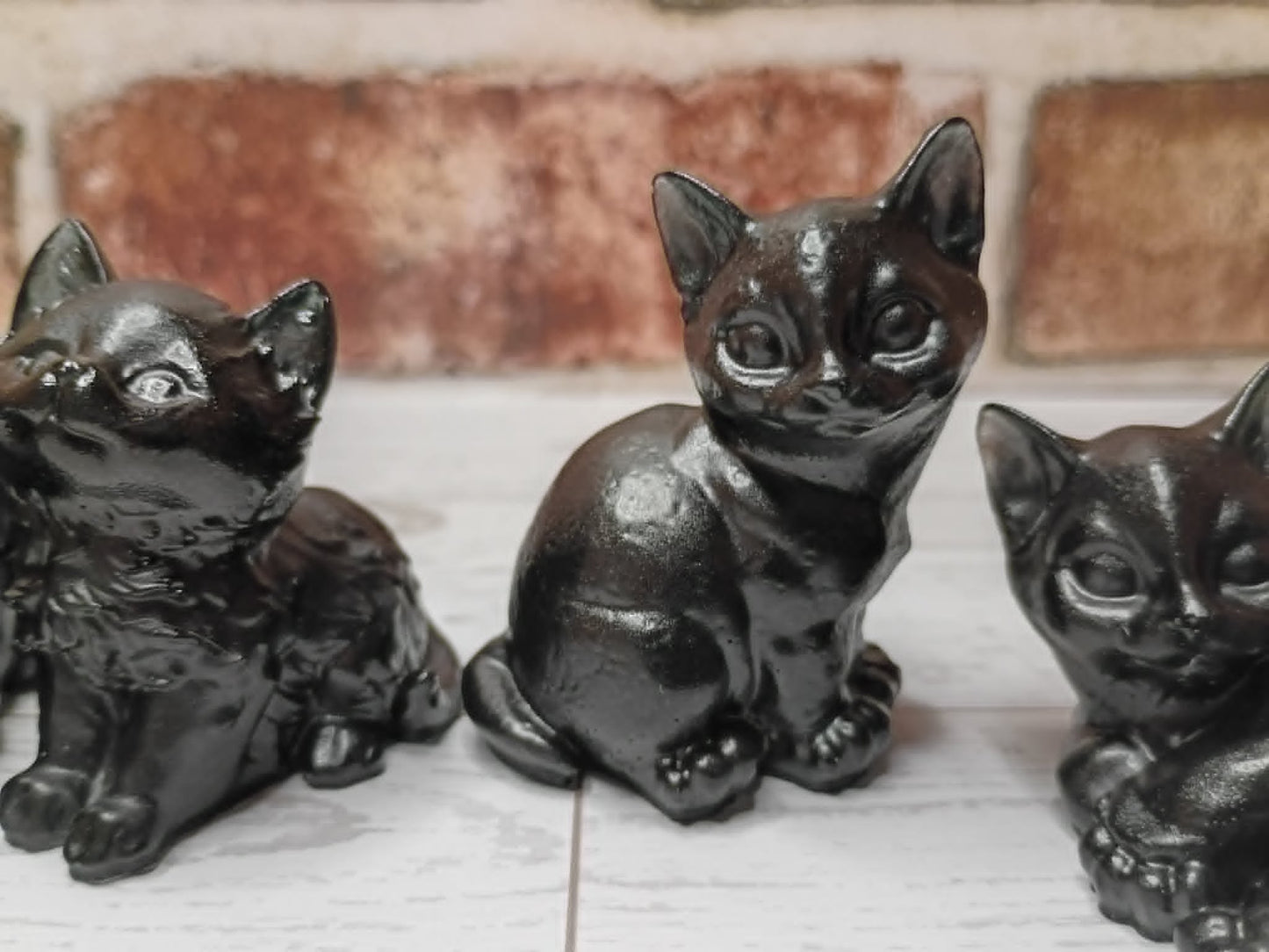 Three glossy black cast-iron kitten figurines displayed on a white wooden surface with a rustic brick wall background.