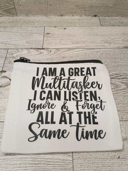 A white canvas zippered pouch with black text that reads 'I AM A GREAT Multitasker I CAN LISTEN, Ignore & Forget ALL AT THE Same time' laid flat on a light wood floor.