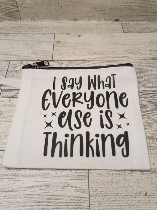 A white canvas zippered pouch with black text reading 'I Say What Everyone Else Is Thinking' and small starburst accents, placed on a light wood floor.