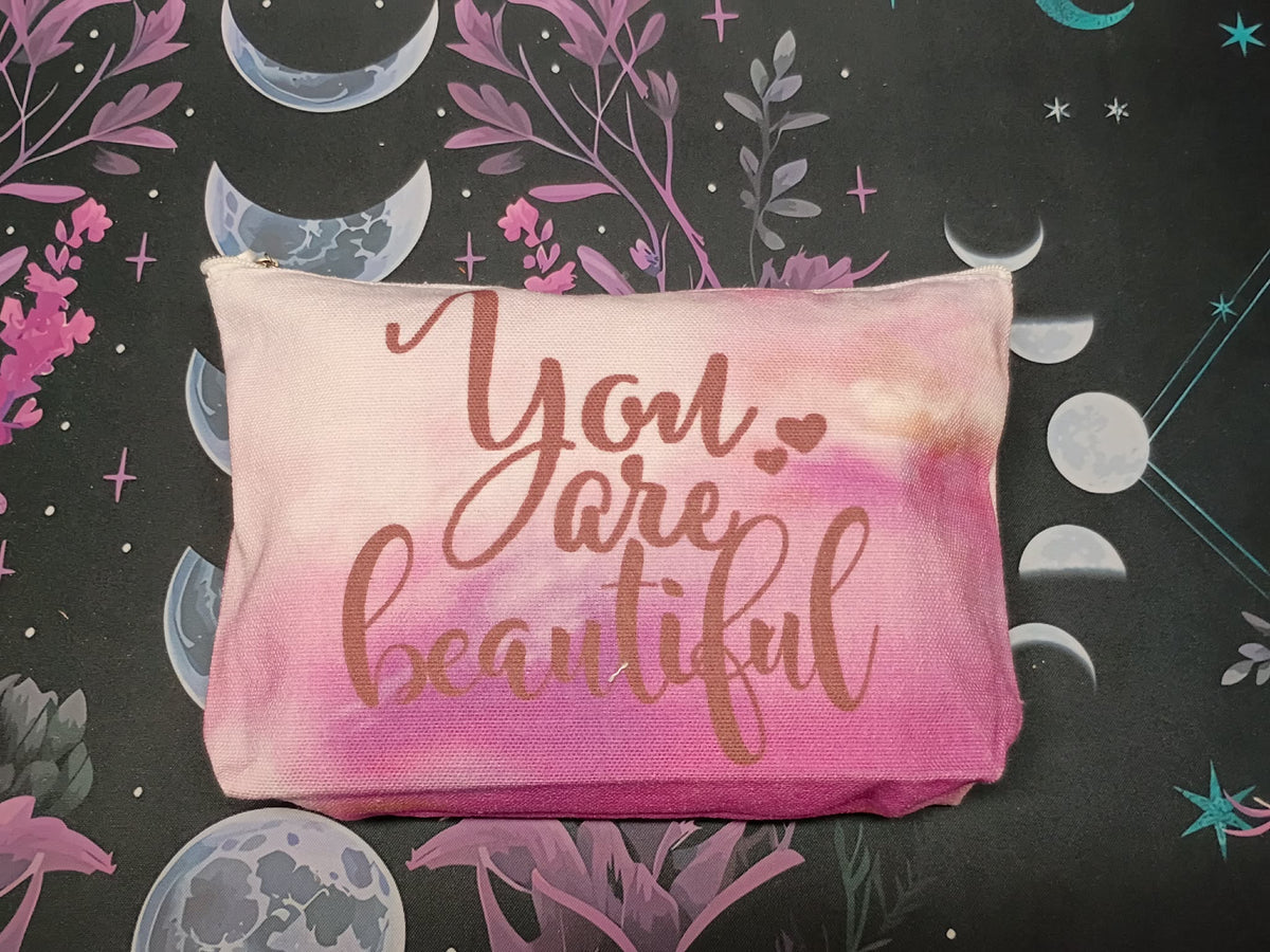 A rectangular fabric pouch with a pink and peach watercolor wash, featuring the phrase 'You are beautiful' in rose gold cursive lettering. The pouch is displayed flat against a dark background decorated with celestial and botanical illustrations.