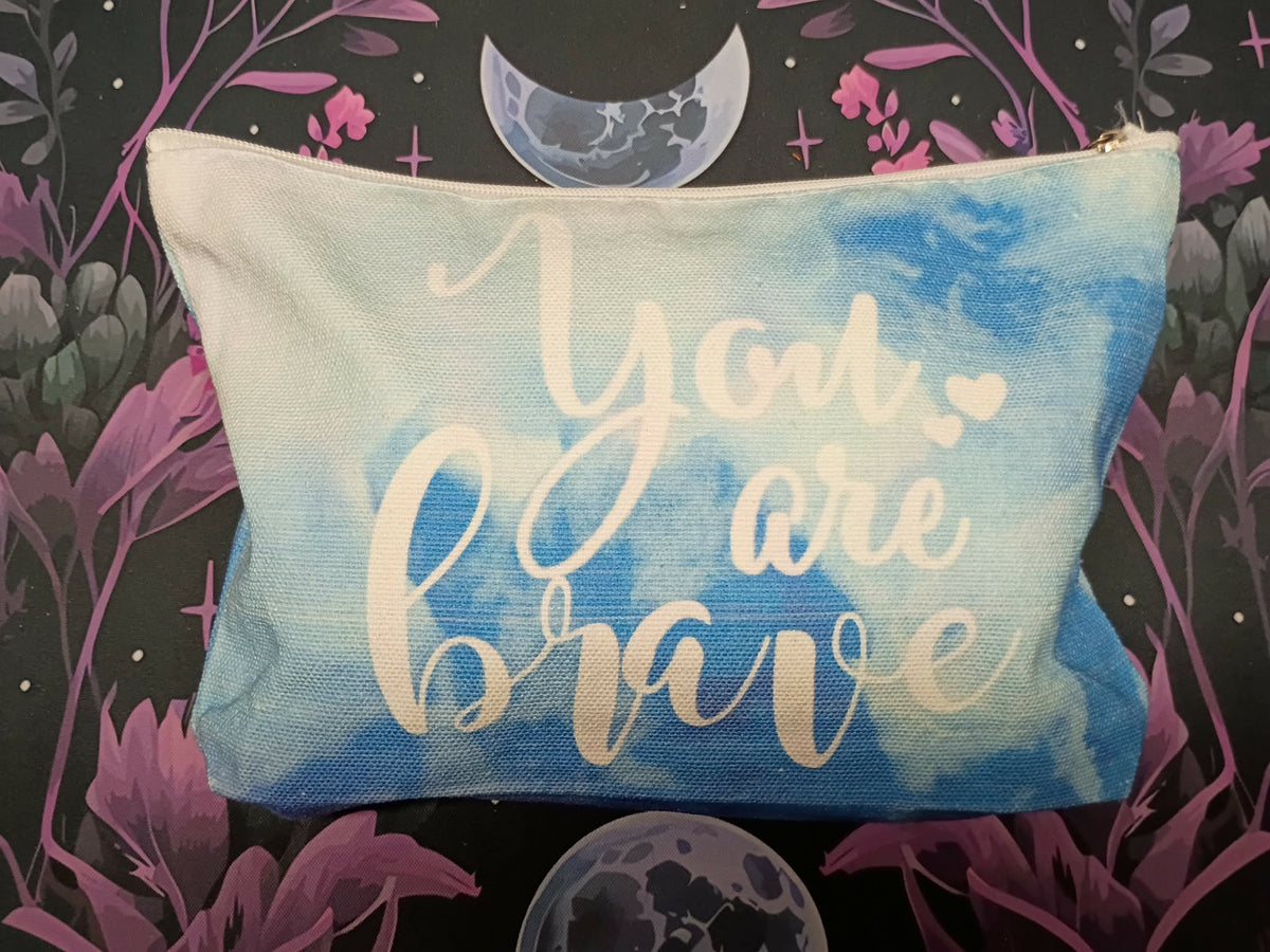 A rectangular canvas pouch with a blue and white watercolor design, featuring the phrase 'You are brave' in white script. The pouch is displayed against a dark background with a purple floral and celestial pattern.
