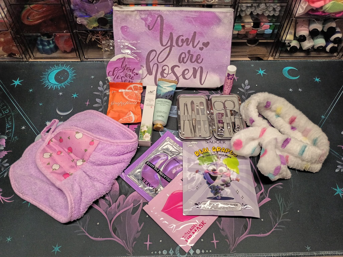 A curated beauty gift set arranged on a dark patterned mat, featuring a lavender zip pouch with 'You are chosen' text, a pink towel pouch with rhinestones, a white fuzzy bunny ear headband, nail care tools in a clear case, and several branded packets including 'Glow Organics' and 'Dr. G's Facial'.