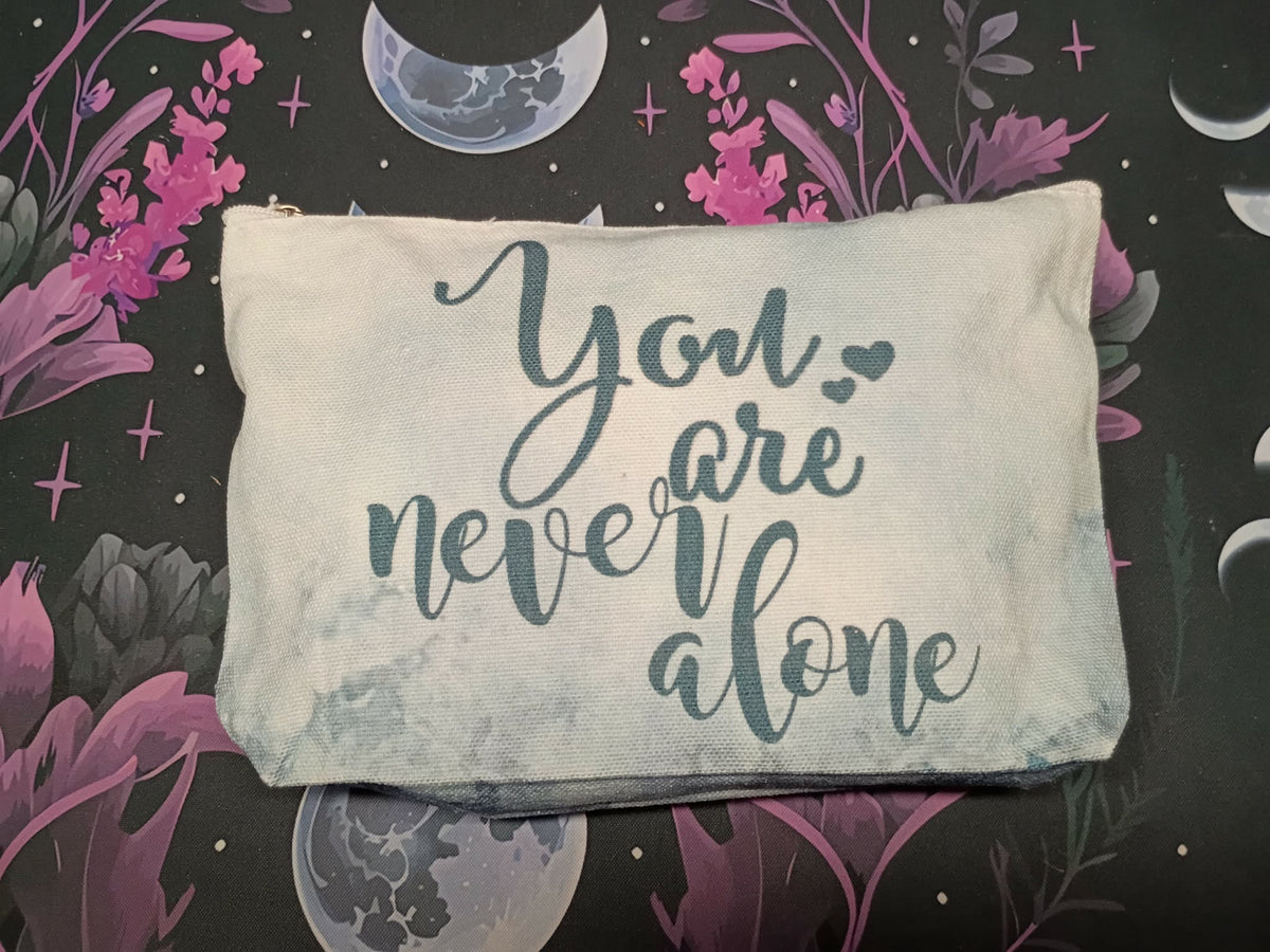 A rectangular canvas pouch with a distressed off-white finish, featuring the phrase 'You are never alone' in dark teal script. The pouch is laid flat on a dark, decorative background with purple floral elements and celestial moon illustrations.