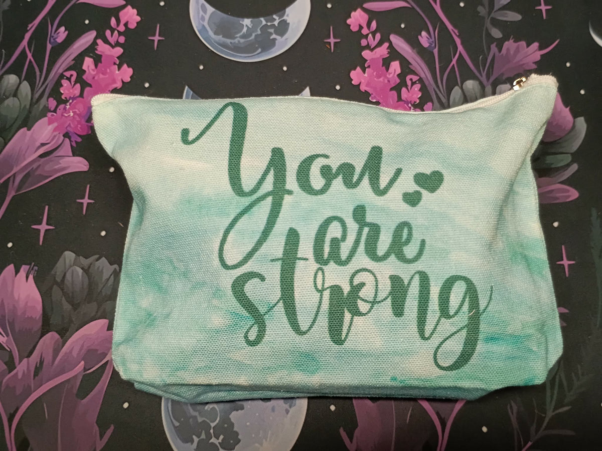 A rectangular zippered pouch with a watercolor mint green and teal wash, featuring the phrase 'You are strong' in dark green script. The pouch is displayed flat against a dark background with a purple and black floral, celestial, and moon pattern.