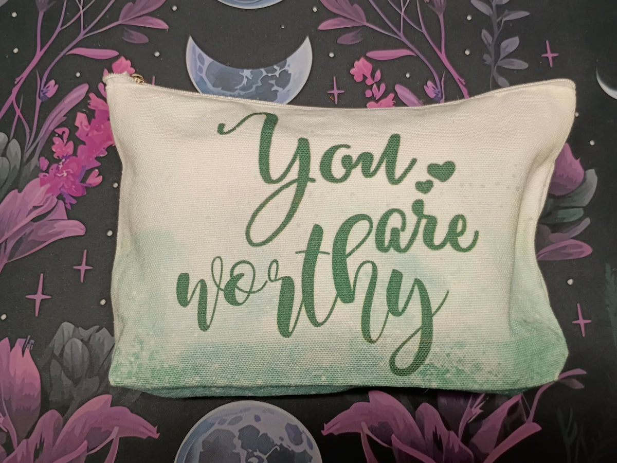 A rectangular canvas pillow with the phrase 'You are worthy' in green script, set against a dark background decorated with purple flowers, moons, and stars.