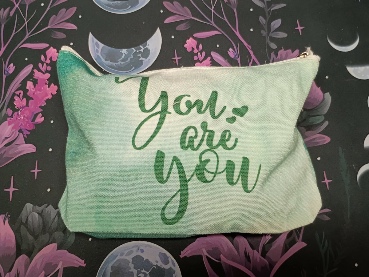 A mint green canvas zippered pouch with the words 'You are you' printed in dark green script, displayed on a dark background with purple flowers and moon illustrations.