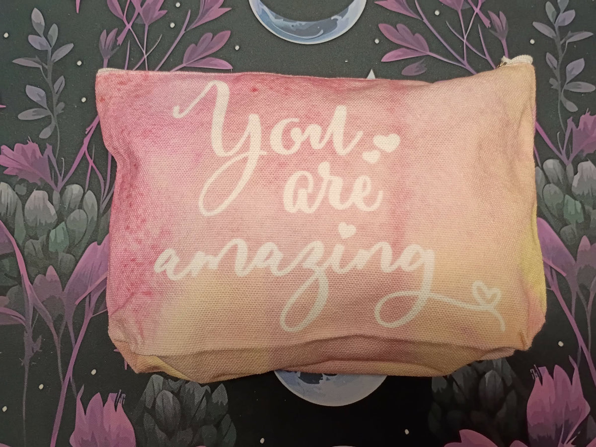 A rectangular zippered pouch with a pink and peach watercolor wash, featuring the phrase 'You are amazing' in white script, set against a dark floral background.